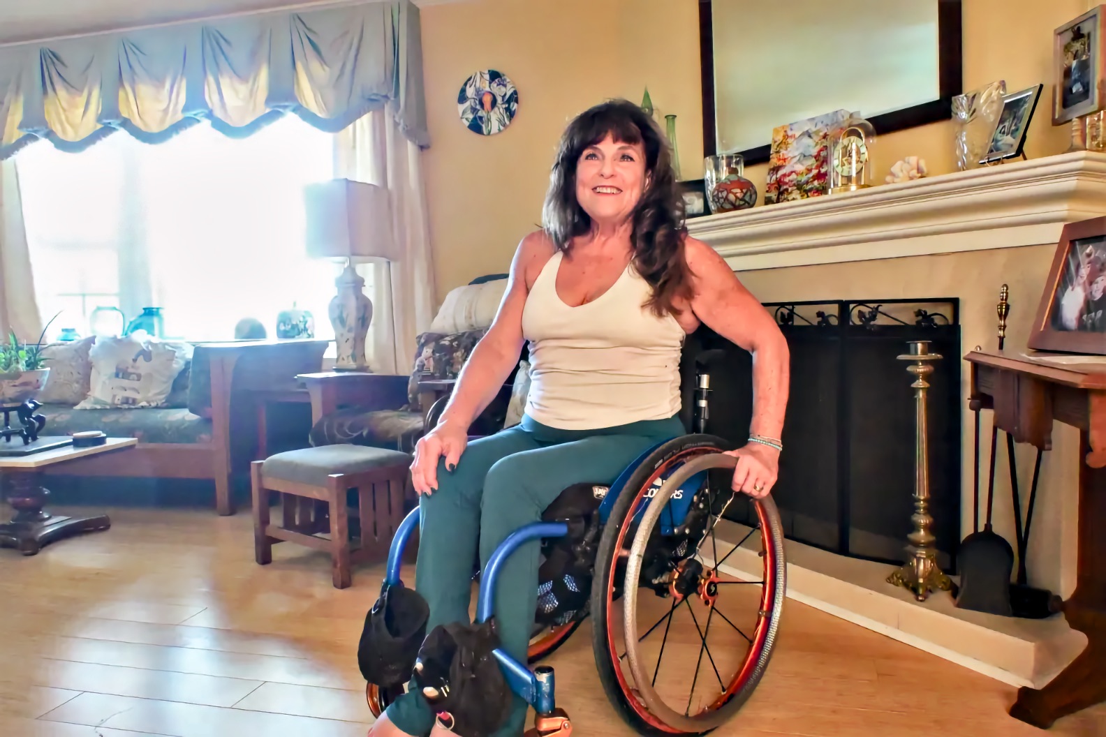 A woman with long dark hair sits in a manual wheelchair with a blue frame and orange rims in a living room, smiling and looking upward. One hand rests on the wheel rim and the other on her leg. Behind her are a fireplace with a white mantel, framed photographs, a mirror, and a curtained window.