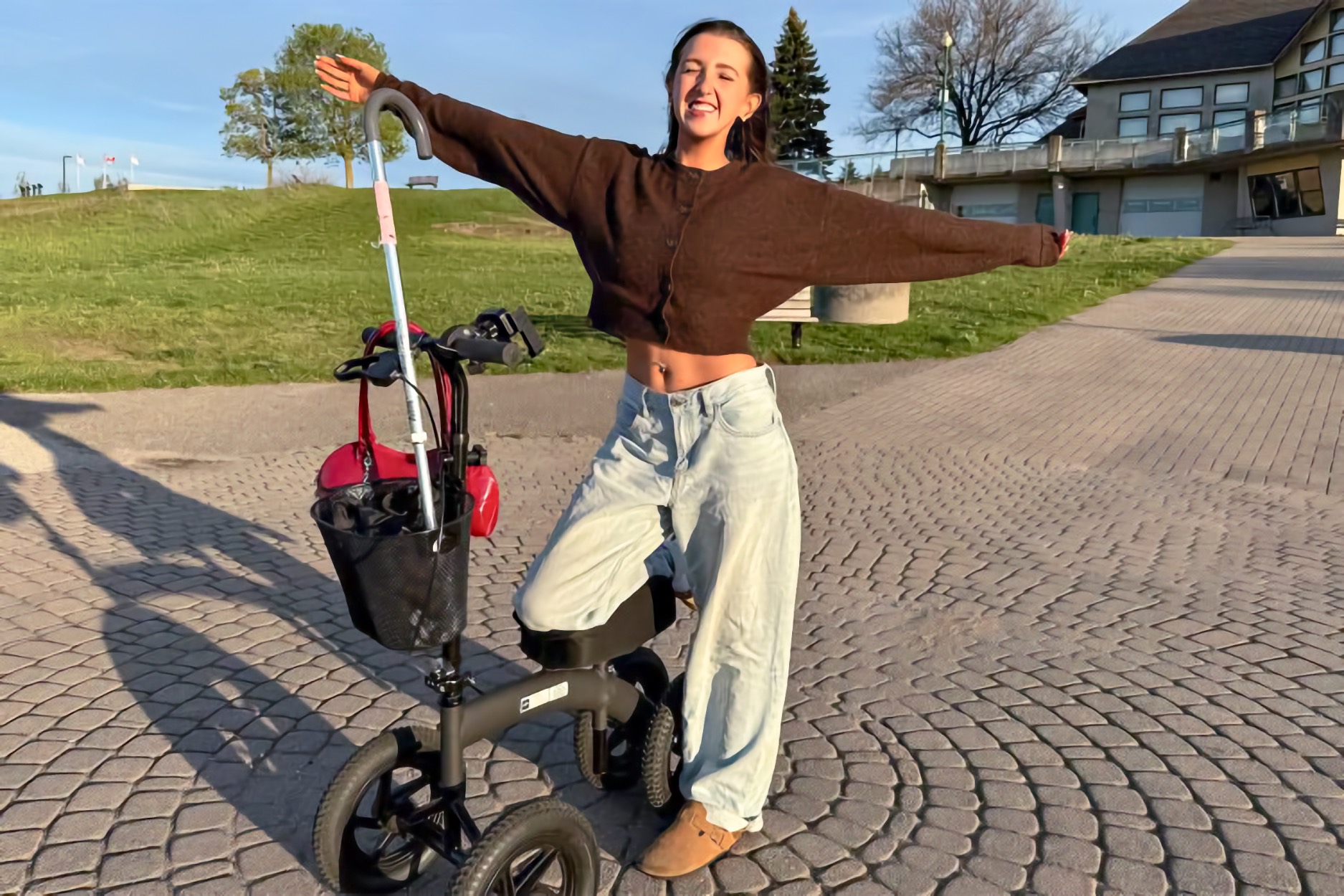 A young woman with a disability stands beside a wheeled knee scooter on a paved outdoor plaza, smiling with her arms stretched wide and one leg bent behind her. The scooter has a handlebar and a small front basket. A grassy hill, trees, and a building stand in the background.