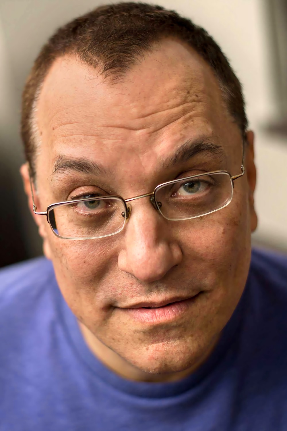 	A man with short dark hair and thin metal framed glasses, photographed closely from the shoulders up, his head tilted slightly as he looks directly at the viewer with a faint smile.
