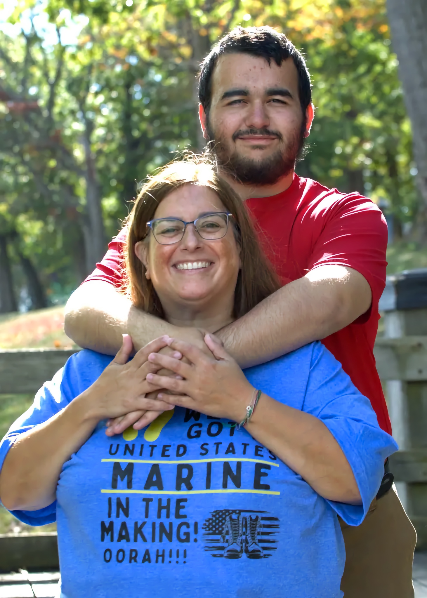 John — a bearded adult man with a language processing disorder — stands close behind his mother, Kristen Boucher, wrapping his arms around her shoulders in an affectionate hug; his proud, tender expression shows clear love and admiration for her as she smiles with calm confidence. Kristen’s T-shirt reads: “We’ve got a United States Marine in the Making! OORAH!!!”