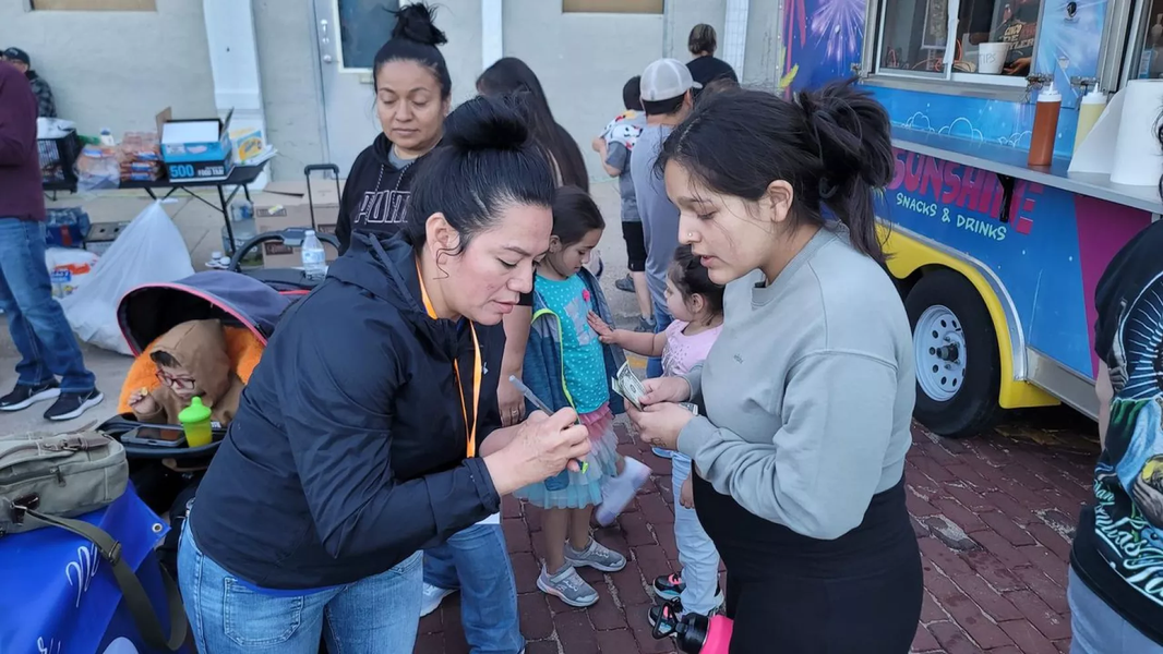 Latina community members gather at an outdoor brick plaza: two adult women engage closely in the foreground, a young child stands between them, a second adult woman stands directly behind them, a toddler stands to the right, and an infant sits in a stroller to the left.