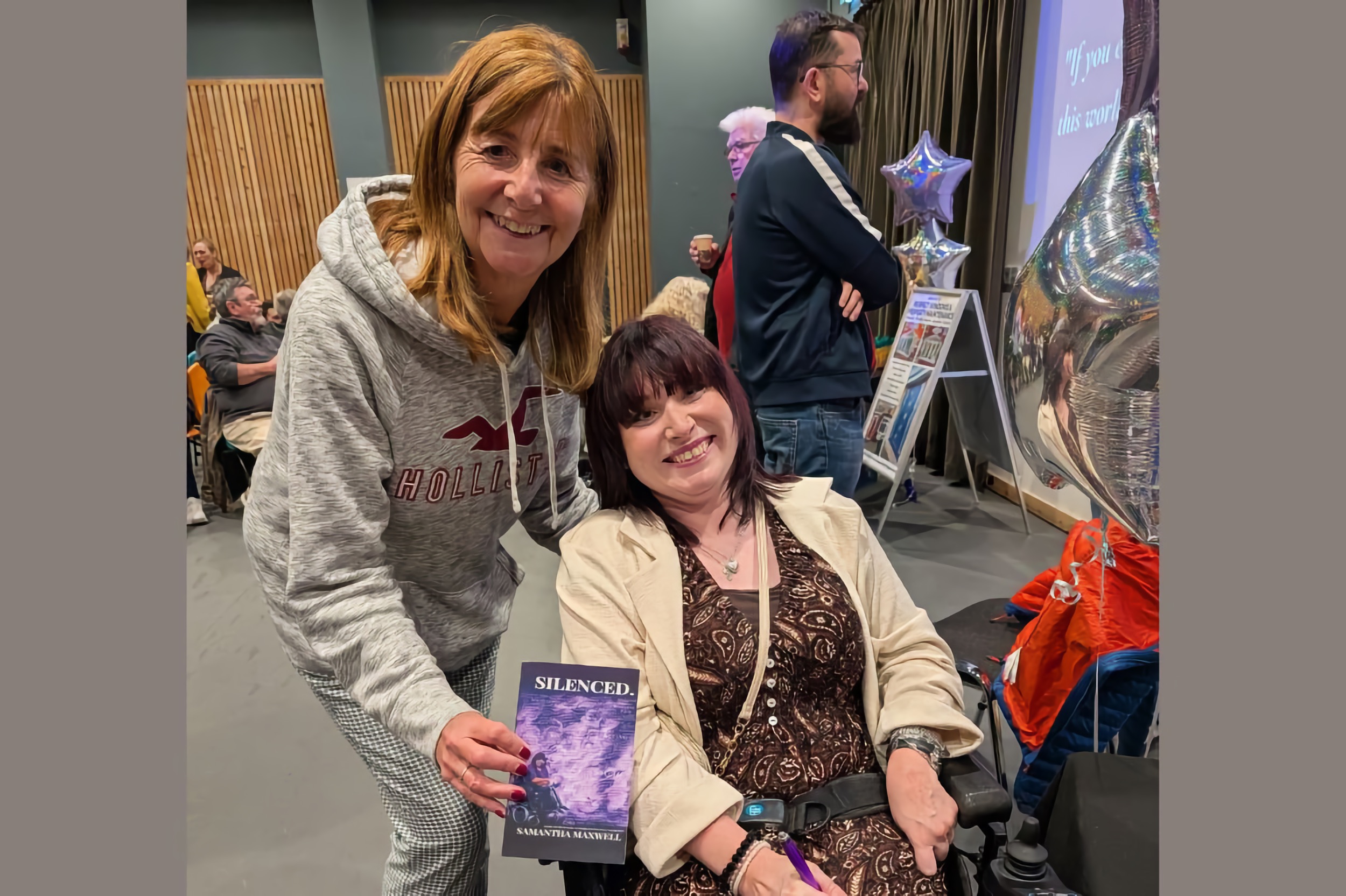 Smiling woman in a grey hoodie stands beside Samantha Maxwell, who is seated in a powered wheelchair and holding a copy of “SILENCED.”; balloons, a projector screen, and an audience are visible in the busy hall.