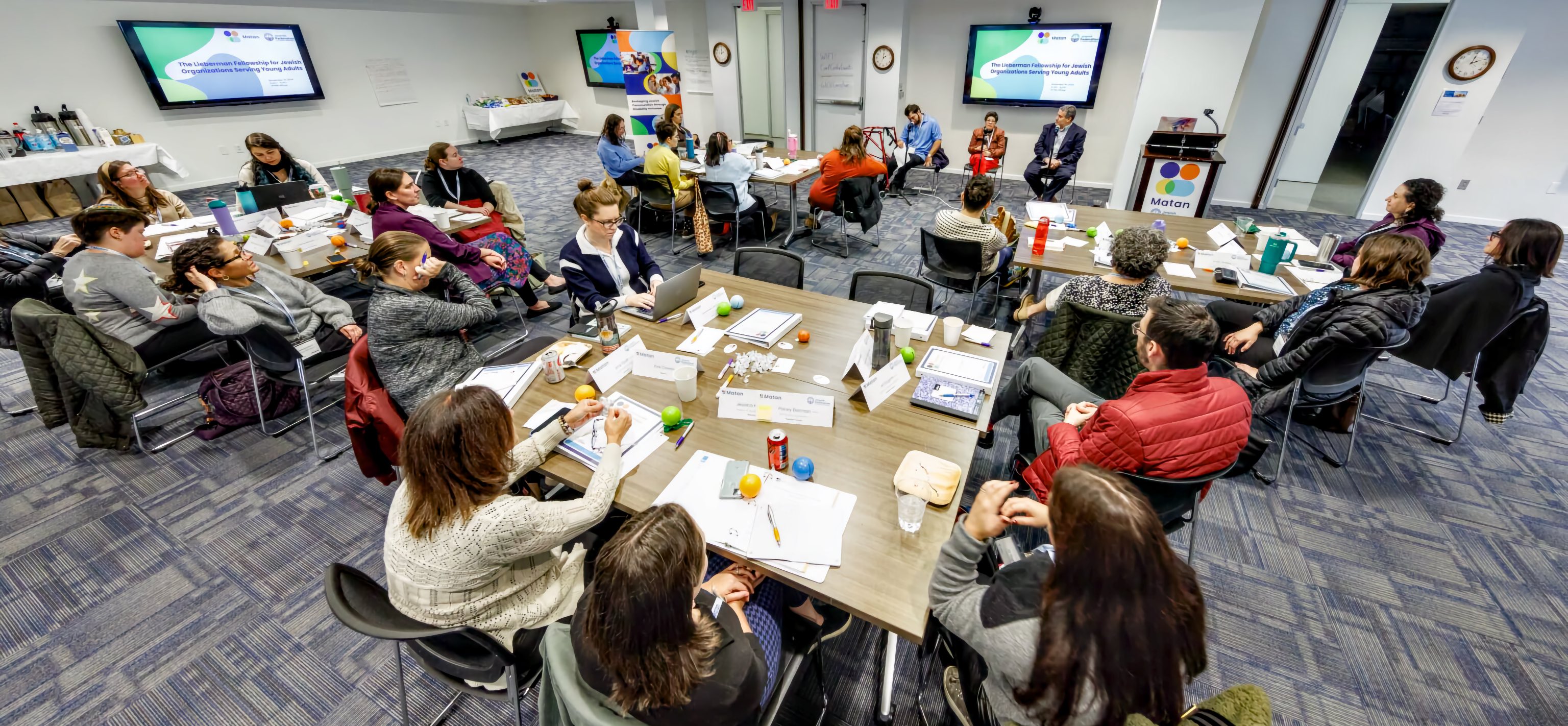 A large group sits at U-shaped tables in a conference room, facing a panel of four speakers near two wall-mounted screens.