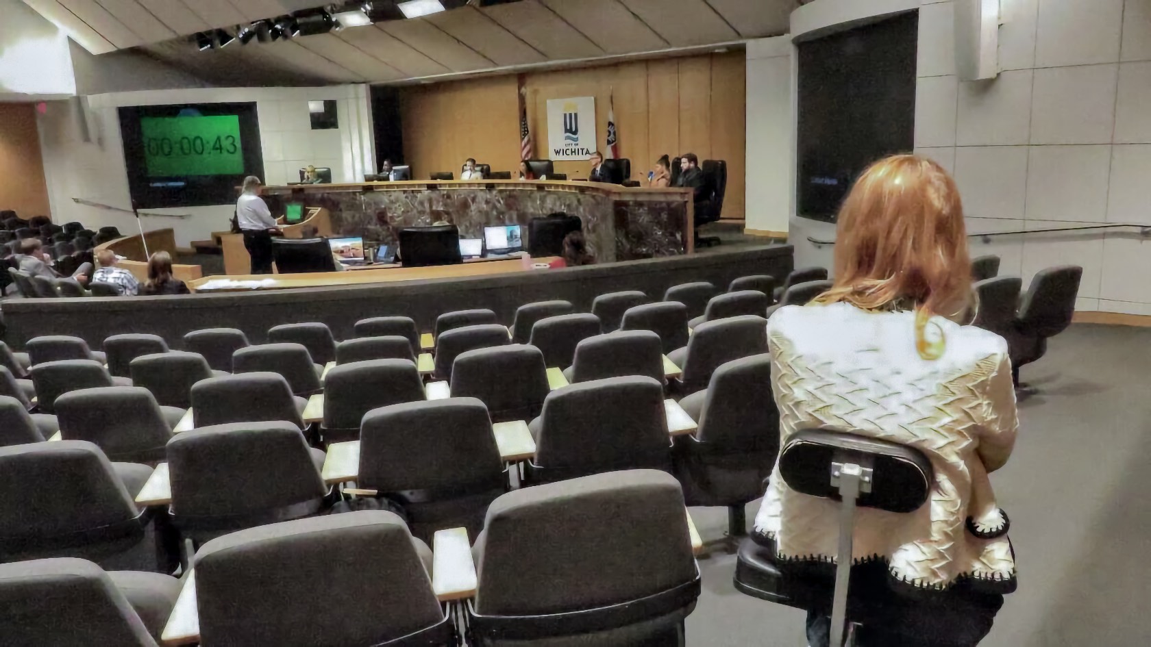 An adult woman with long red hair, seated in a power wheelchair, viewed from behind at the rear of a public hearing chamber. Many rows of empty gray seats with folding writing surfaces separate her from the front of the room. Ahead, several adults are seated behind a raised marble bench beneath a large city seal, and two flags stand beside them. A man stands at a podium facing the bench. A timer on a screen at the left reads 00:00:43 in green digits.