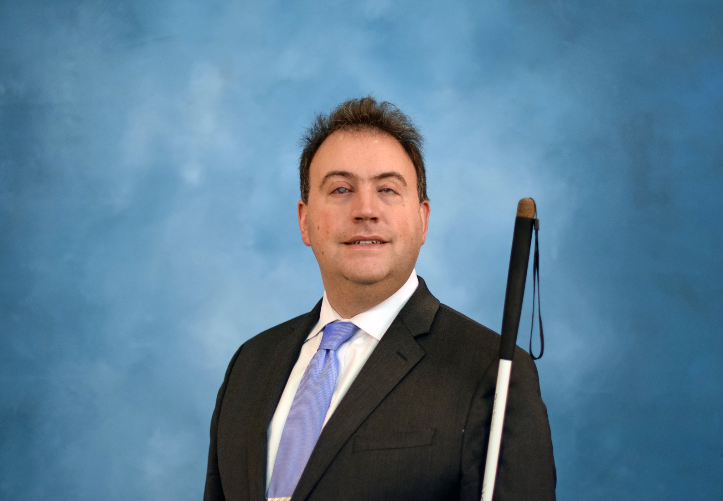 A Blind adult man smiles slightly while holding a long white cane upright beside his shoulder against a blue studio backdrop.