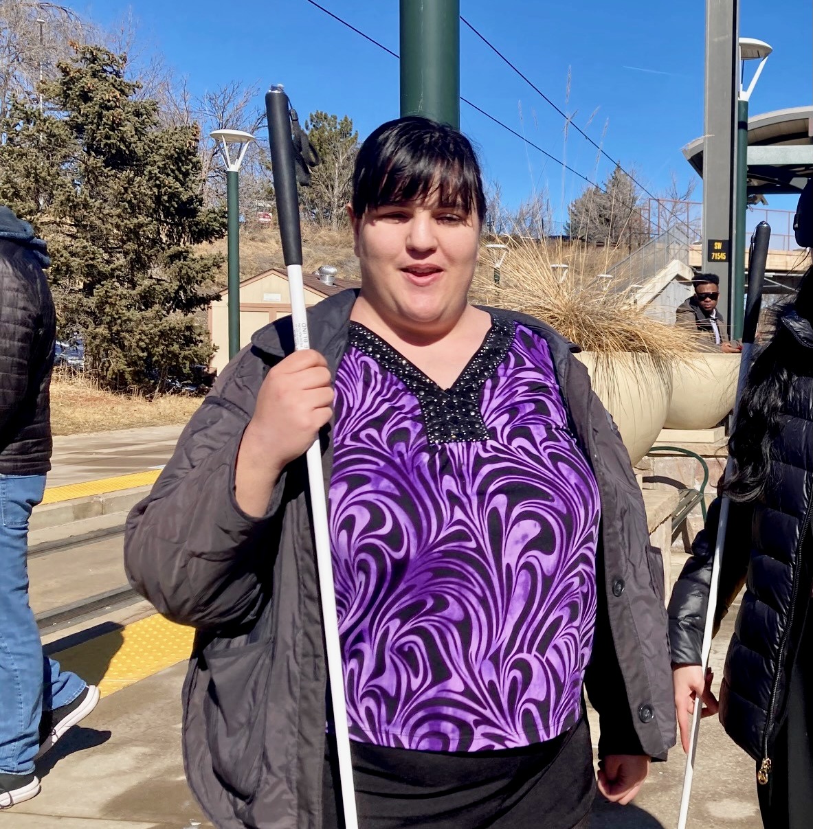 Blind woman in a purple patterned blouse stands outdoors holding a white cane at a train station on a sunny day.