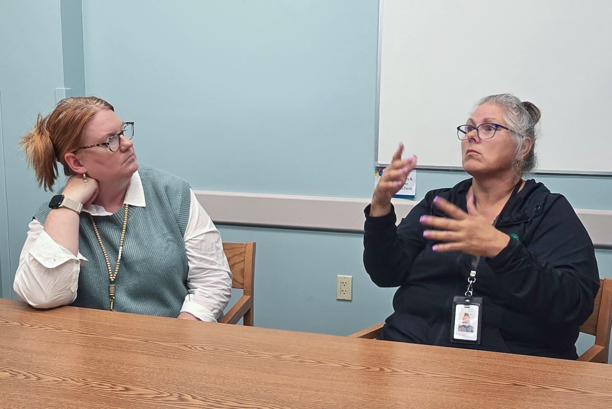 Two adult women at a meeting table; the person on the right signs in ASL while speaking, the person on the left watches.