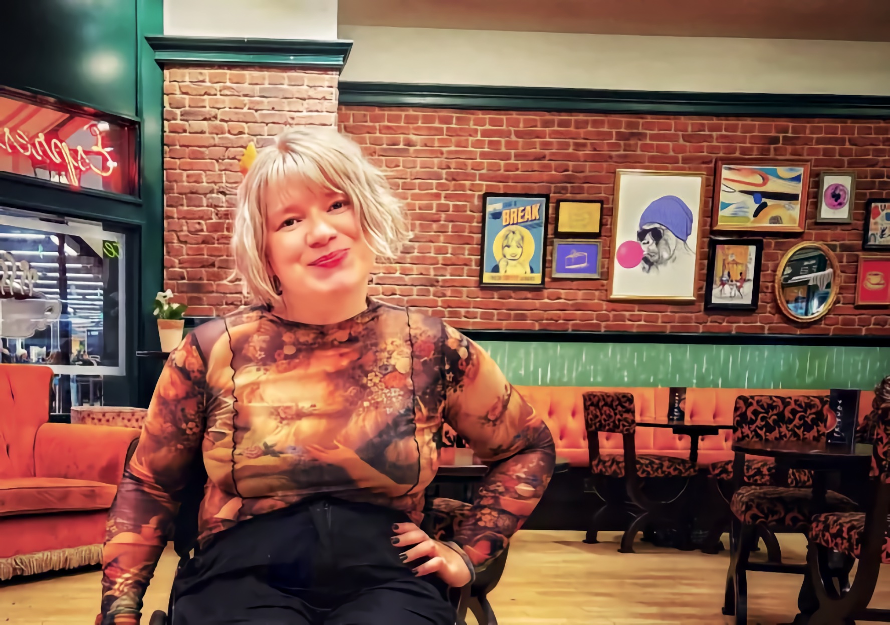 Melissa Parker, a young woman with short platinum blonde hair, seated in a manual wheelchair and smiling in a colorful café with exposed brick walls and framed artwork behind her.