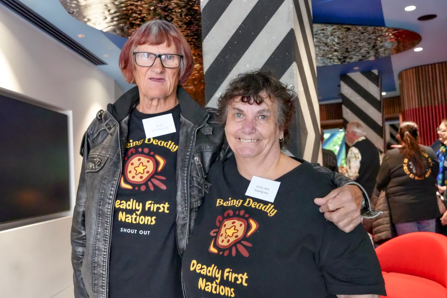 Two First Nations women—Aunty Jane Rosengrave (Yorta Yorta, senior adult) and Sarah Waardenburg (Gunaikurnai, adult)—stand together at an indoor launch event, smiling toward the camera; both wear black “Being Deadly — Deadly First Nations Shout Out” shirts and name tags. Other attendees are blurred behind them.
