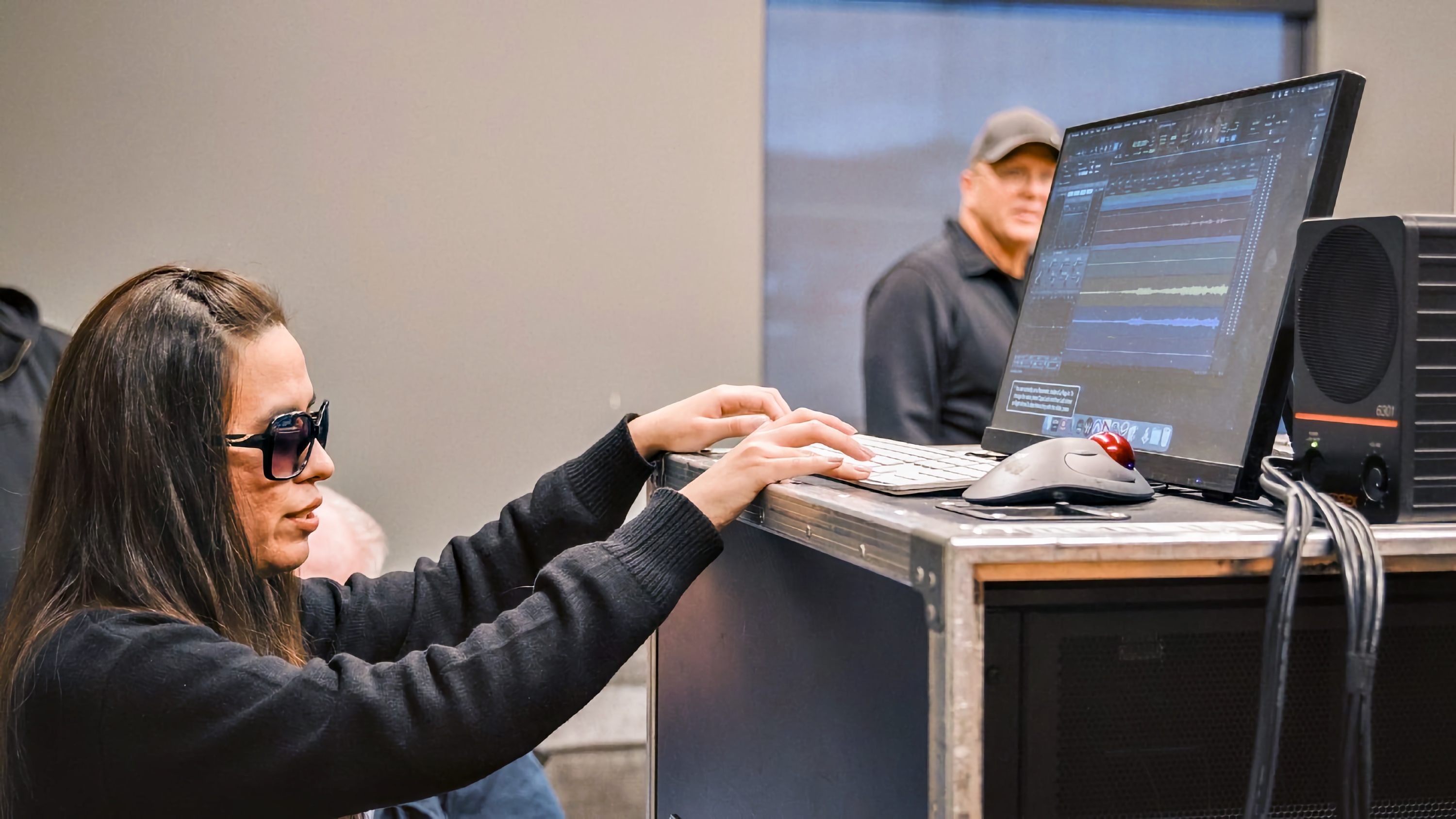 Blind woman uses keyboard to edit audio waveforms on screen in a professional studio setup with speaker and production case nearby.