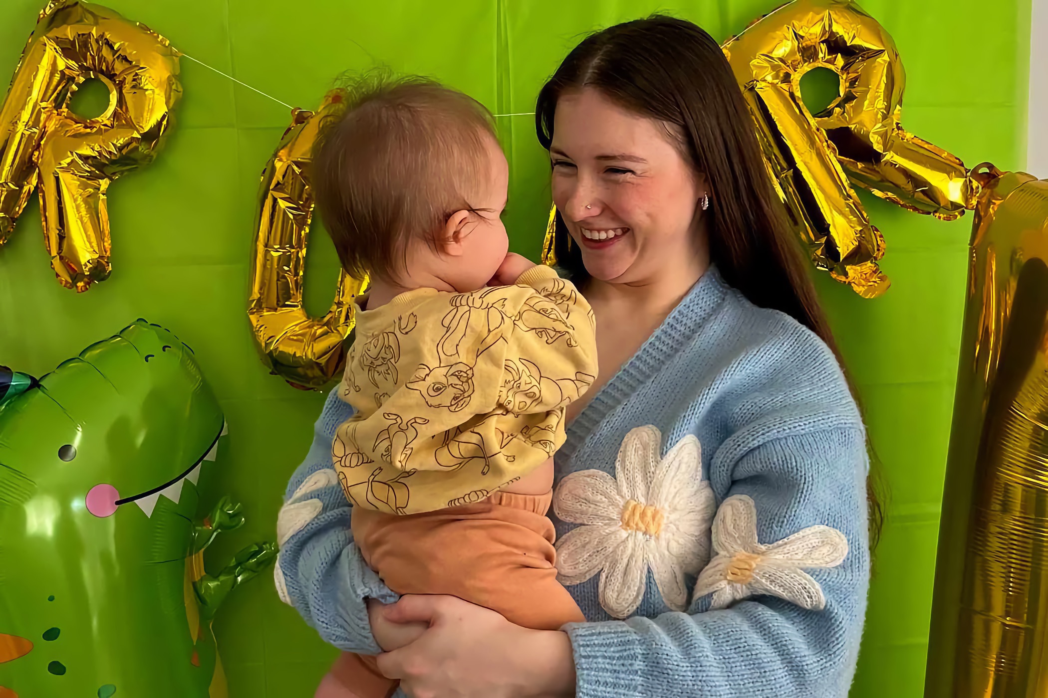 Woman in blue sweater smiles at baby in front of green backdrop with gold letter balloons and a dinosaur balloon.