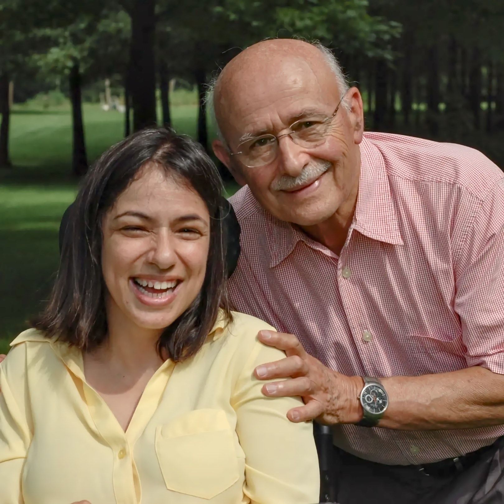 A young adult Middle Eastern woman with dark shoulder length hair smiles widely, seated in a power wheelchair with a black headrest visible behind her shoulder. An older Middle Eastern man with glasses and a white mustache leans in beside her, resting his hand on her shoulder and smiling. They are outdoors in a park with trees and a green lawn behind them.