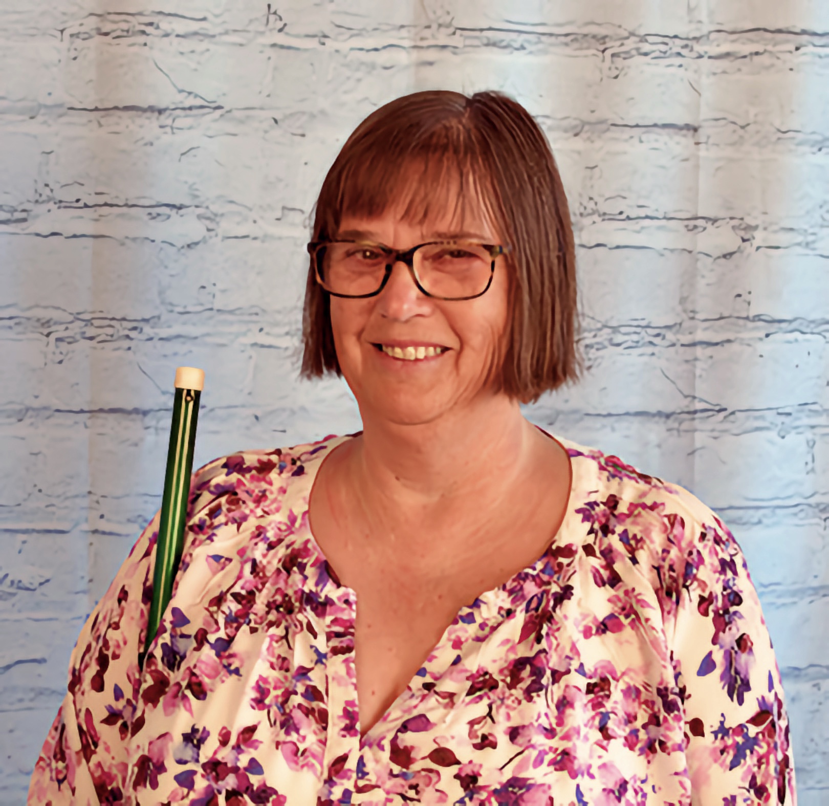 A middle-aged woman with chin-length brown hair and glasses holds a green pencil upright beside her smiling face.