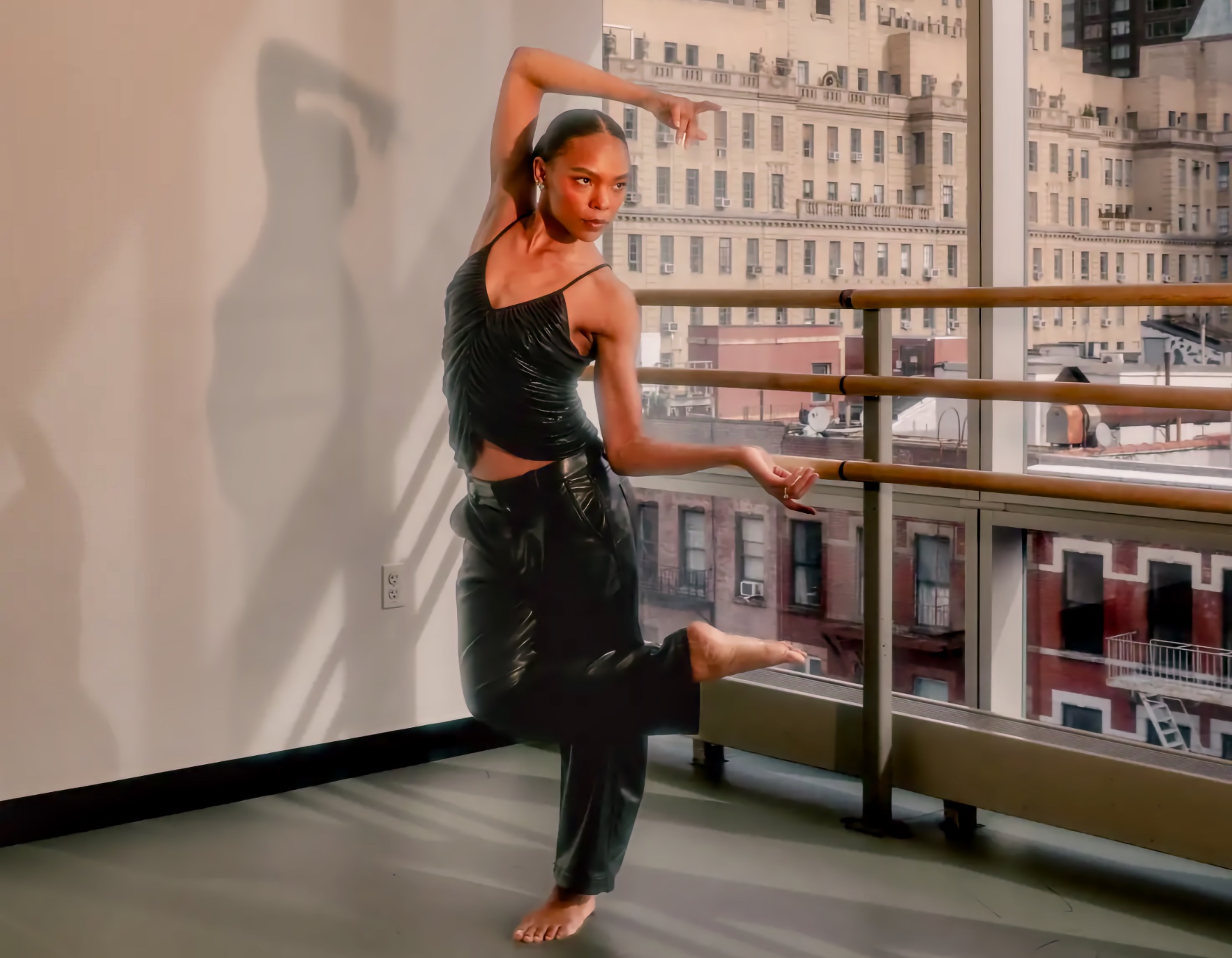 Young adult Black woman dancer Samantha Figgins, who has hearing loss, balances barefoot on one leg beside ballet barres in a sunlit studio, with one arm curved overhead, one hand extended, and tall windows opening onto city buildings behind her.