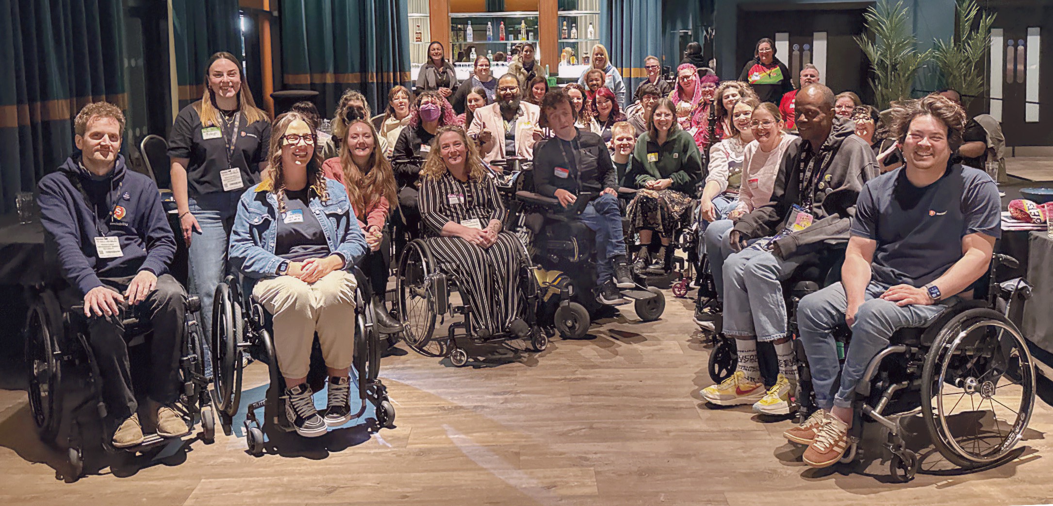 A group of approximately 35 men and women ranging from young adults to adults poses for a photograph in a dimly lit event venue with teal curtains and circular pendant lights. Multiple people use manual wheelchairs, with several wheelchair users prominent in the foreground. A Black man stands at the front right of the group. Hardwood floors and a bar area are visible in the background.