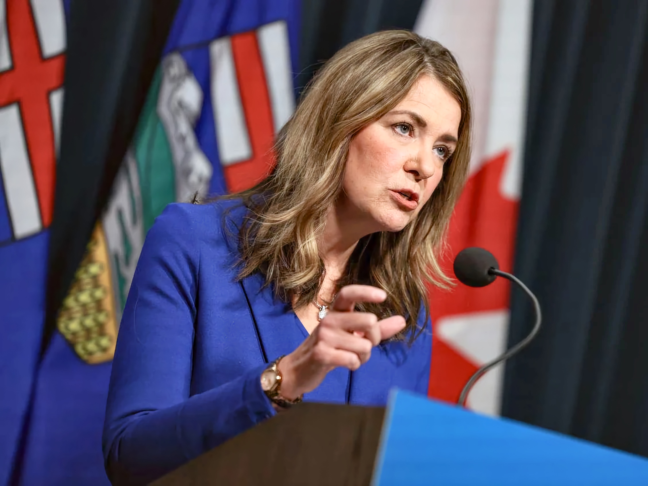 A woman in her mid-forties with shoulder length brown hair, wearing a blue blazer, speaks at a microphone stand and gestures with her right hand. Behind her, partial views of the Alberta flag and Canadian flag are visible in a formal indoor setting.
