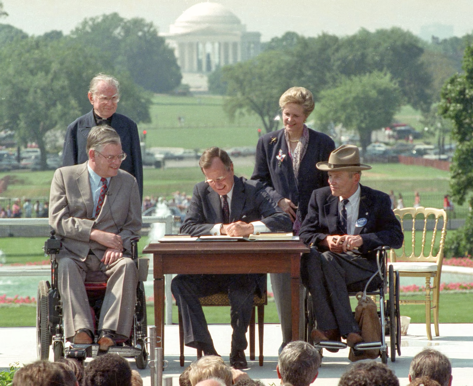 President George H.W. Bush, a male in his mid-60s, sits at a signing table on the South Lawn of the White House. To his left stand Evan Kemp, a male in his mid-50s who uses a wheelchair due to a neuromuscular disability, and Rev. Harold Wilke, a male in his mid-70s who was born without arms. To his right stand Sandra Parrino, a female in her early 50s, and Justin Dart, a male in his late 50s who uses a wheelchair. Bush is signing the Americans with Disabilities Act into law, July 26, 1990.