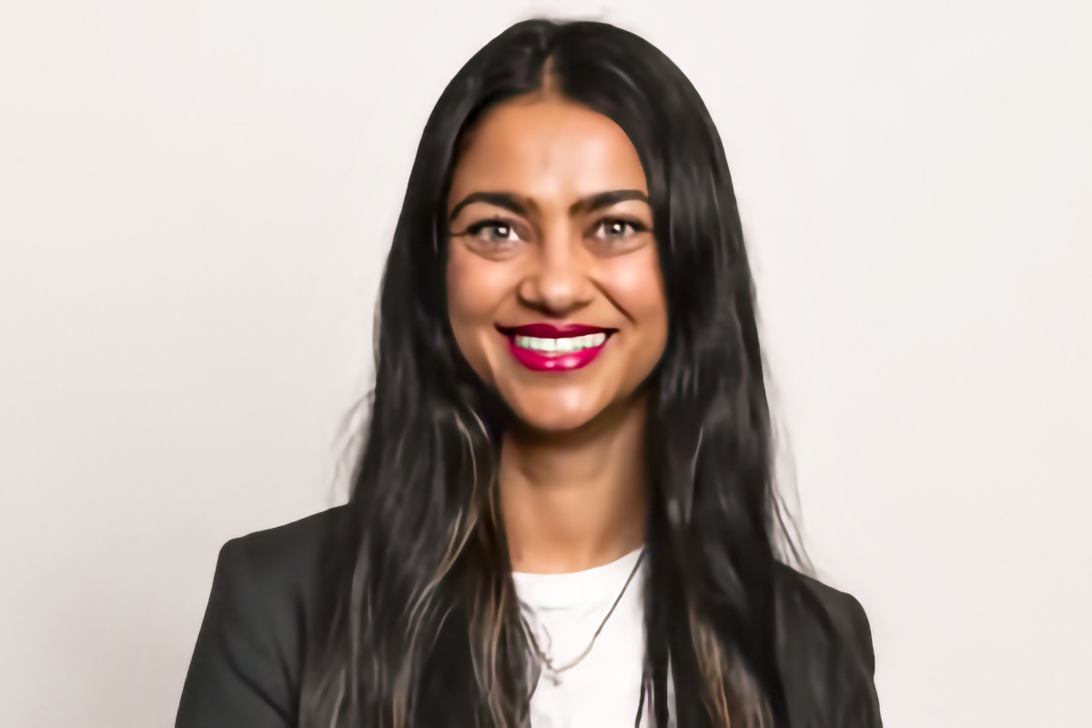 Smiling South Asian adult woman with long dark hair, facing the camera against a plain light background.