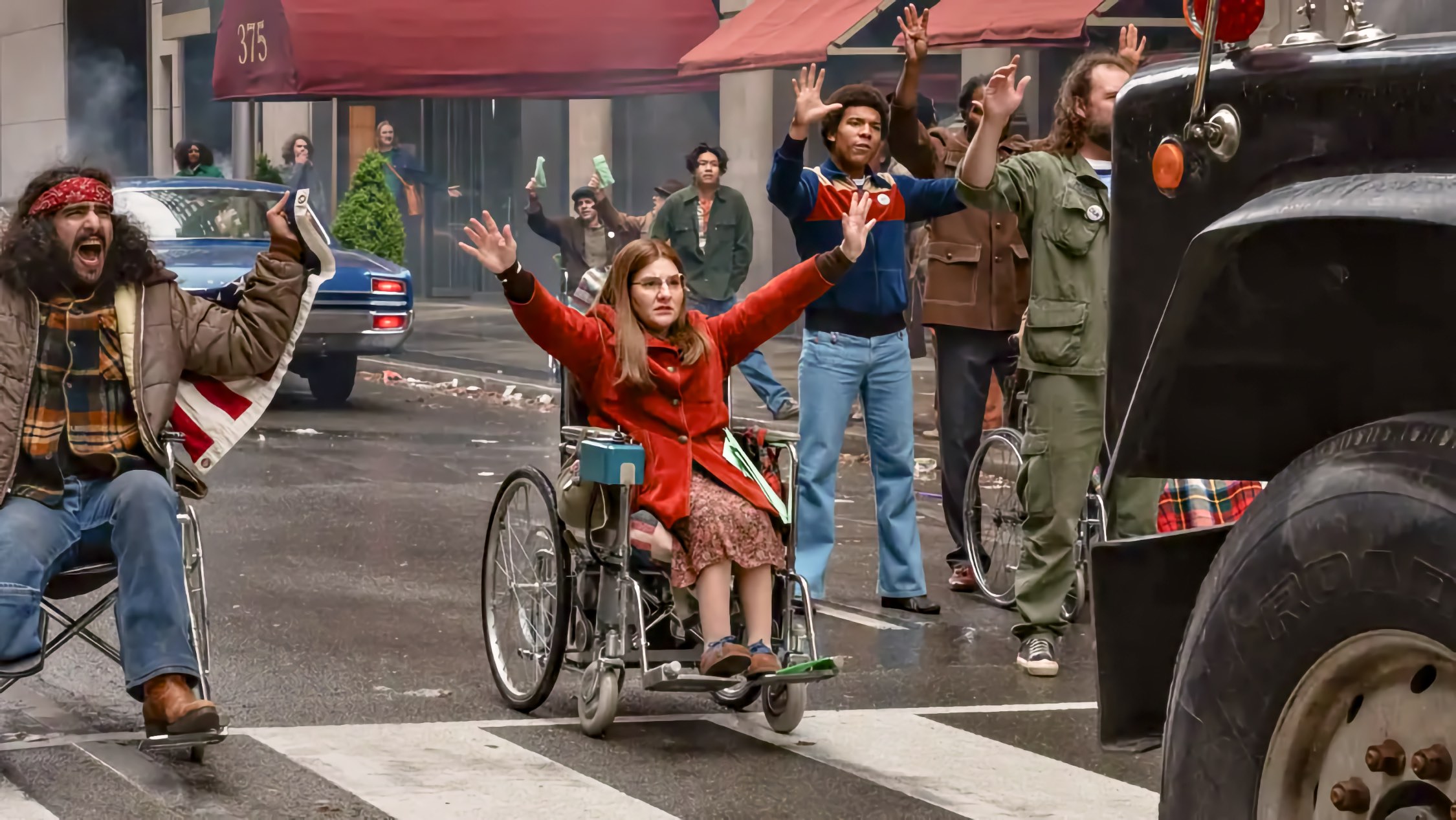 In a city crosswalk, a woman using a wheelchair raises her arms before a truck. A shouting man using a wheelchair and a Black man with raised hands are nearby.
