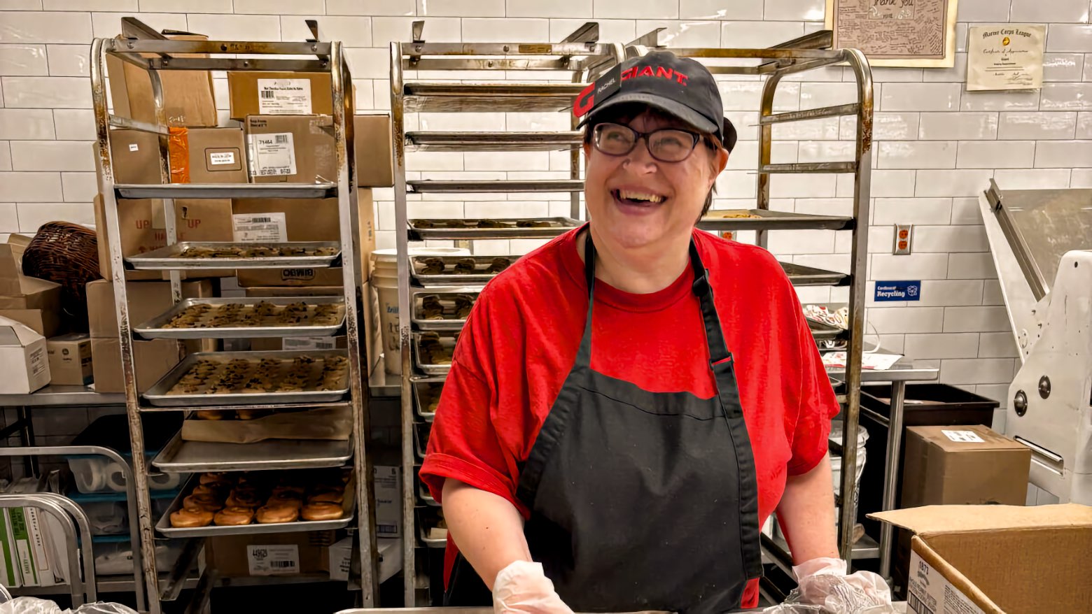 A woman wearing glasses smiles broadly while standing at a bakery work counter, her hands in clear food service gloves. Behind her, tall metal racks hold sheet pans of unbaked cookies and trays of glazed doughnuts.
