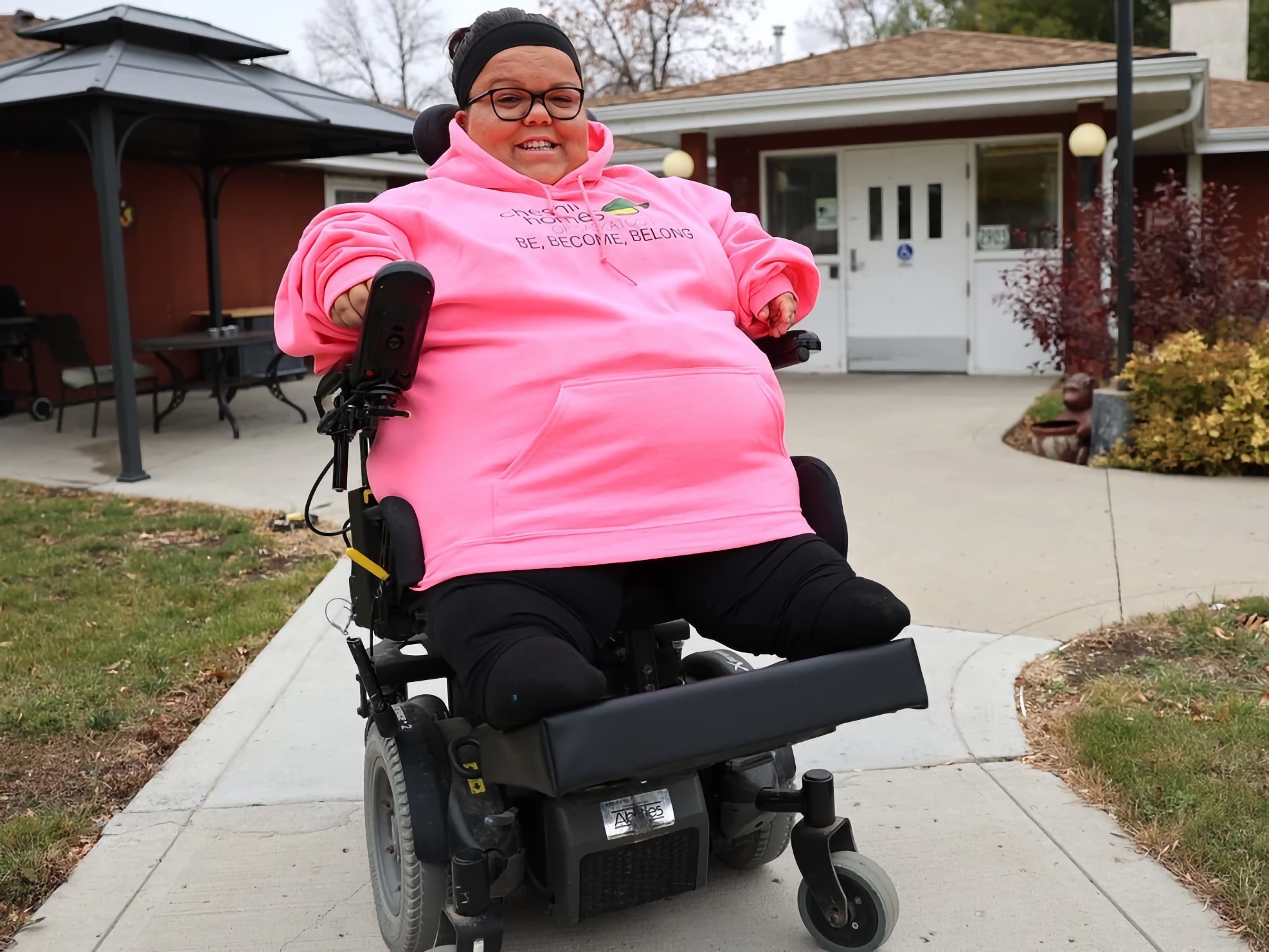 Light-brown–skinned adult woman using a power wheelchair—left hand on a left-mounted joystick, feet on an elevated footboard—smiling on a curved concrete path toward a step-free entrance; gazebo with tables at left, late-season shrubs and patchy grass along the walkway.