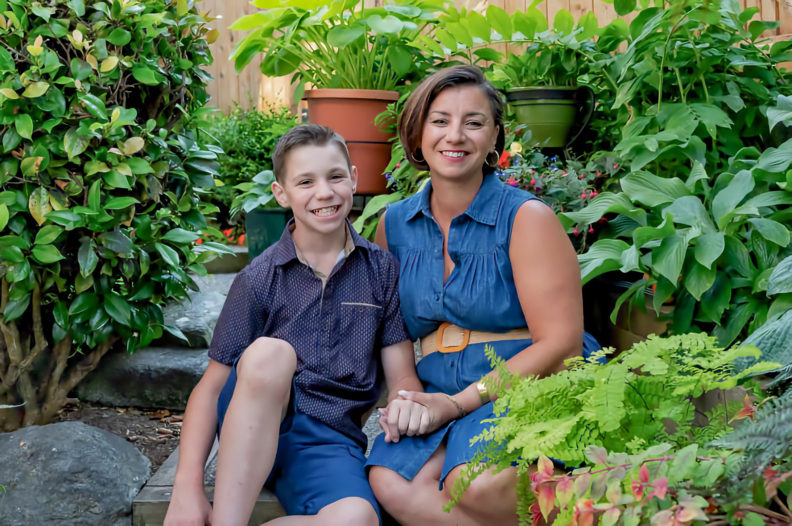 A smiling boy, a child, and his smiling mother sit side by side on garden steps, holding hands, among leafy green plants.