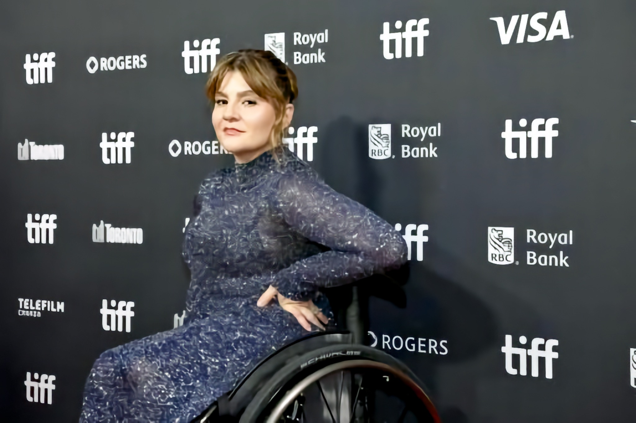 A Woman who uses a manual Wheelchair looks toward the camera in front of a backdrop of Toronto International Film Festival sponsor logos.