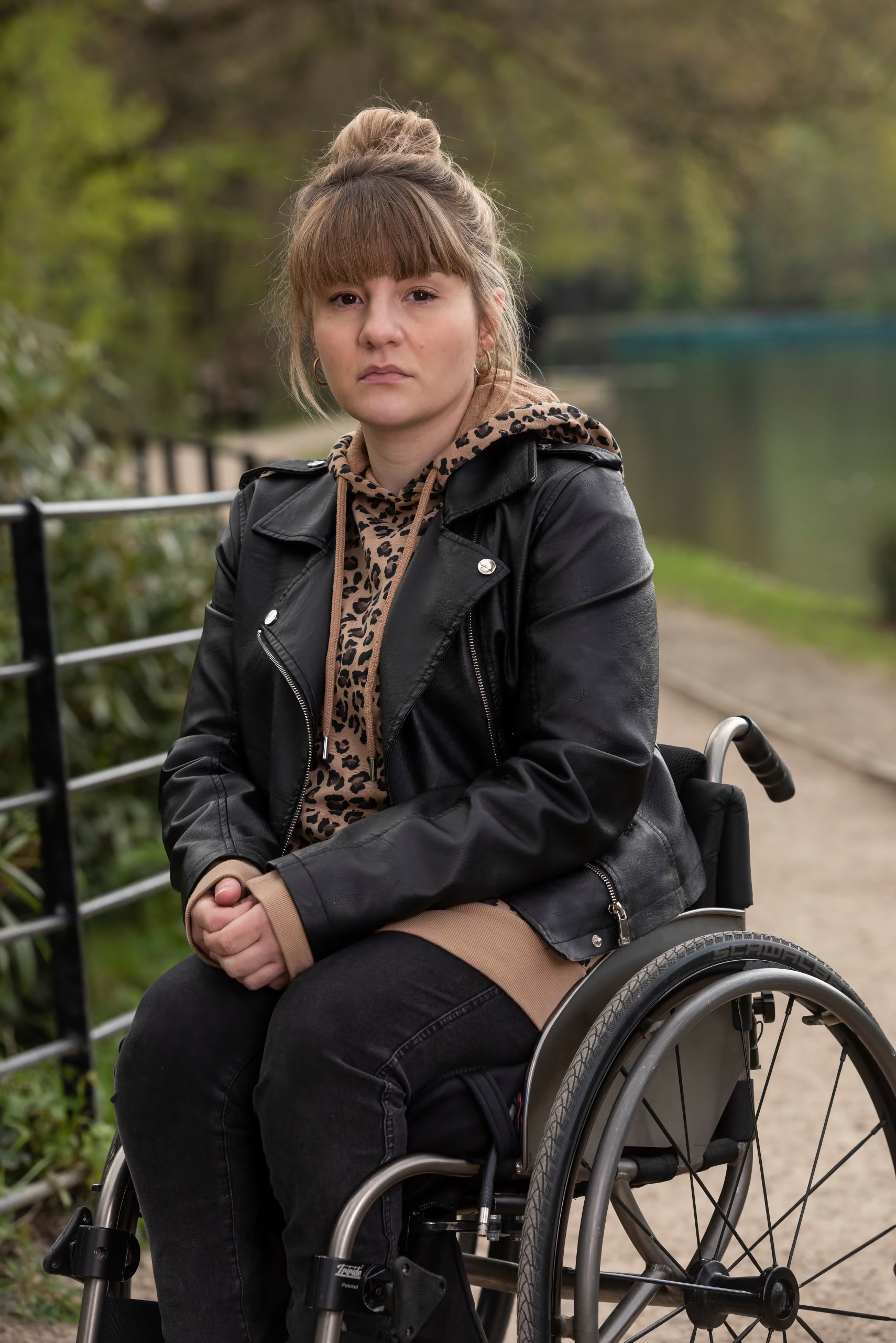 Woman with a high bun wearing a leather jacket and leopard-print hoodie sits in a manual wheelchair beside a calm, tree-lined waterway.