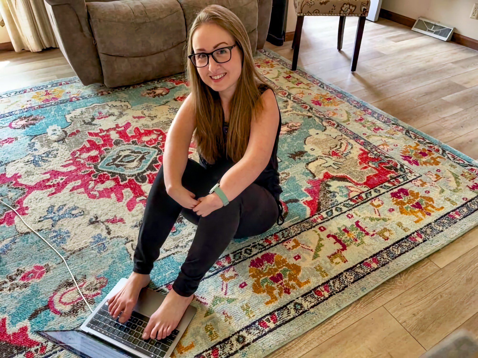 A young woman with long light brown hair and glasses sits cross-legged on a colorful patterned area rug in a home living room. She uses her bare feet to type on an open laptop positioned on the floor in front of her. She wears black athletic clothing and a light-colored smartwatch. An upholstered brown sofa and armchair and hardwood floors are visible in the background.