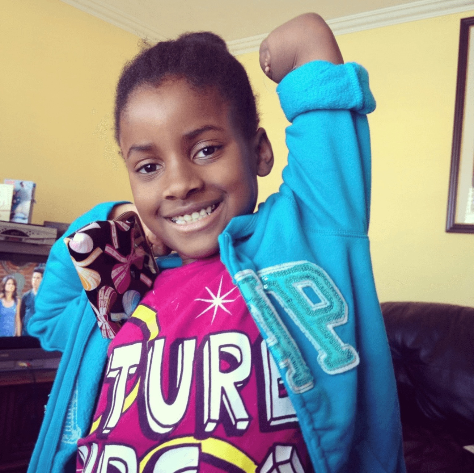 Girl missing her left hand smiles; right arm raised; teal jacket over pink T-shirt; patterned pouch at side; living room.