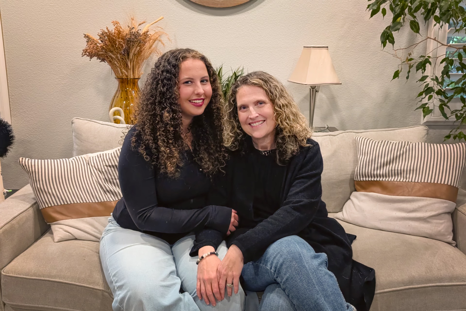 A teenage girl with long curly brown hair and a woman with shoulder-length curly blonde hair sit close together, both smiling, on a sofa.