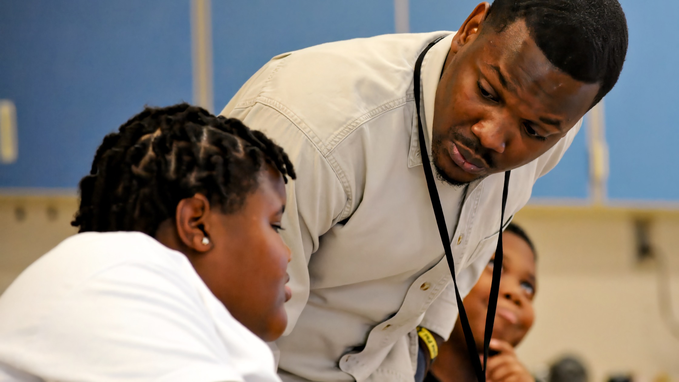 A Black adult male teacher leans forward over two Black elementary school age students seated at a classroom desk, speaking to them at close range while they look down at their work. A blue wall is visible in the background.