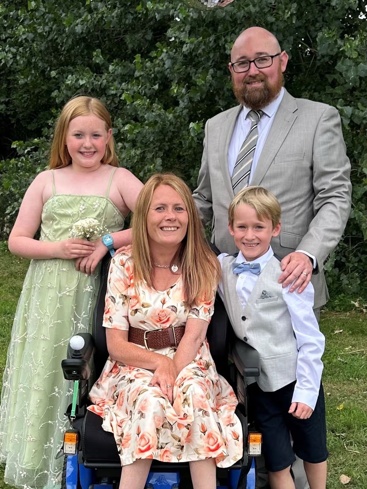 A middle-aged woman seated in a power wheelchair is at center of a family group outdoors. An adult man in his late 30s stands at her side. A young girl of approximately 11 years and a boy child of approximately 9 years complete the group. All four are smiling together.