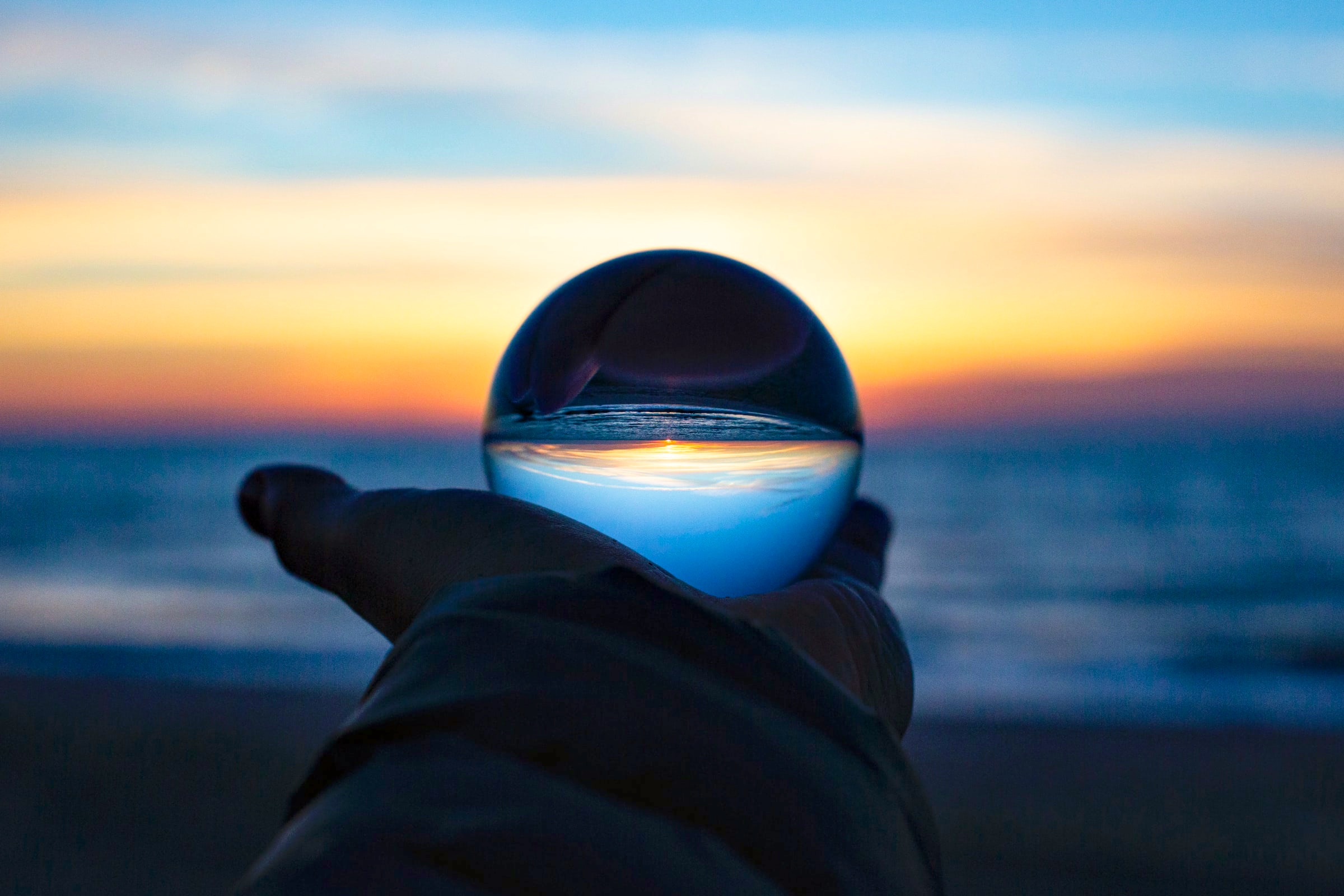 A palm in a dark sleeve holds a crystal sphere that flips a shoreline and horizon against a blurred twilight sky.