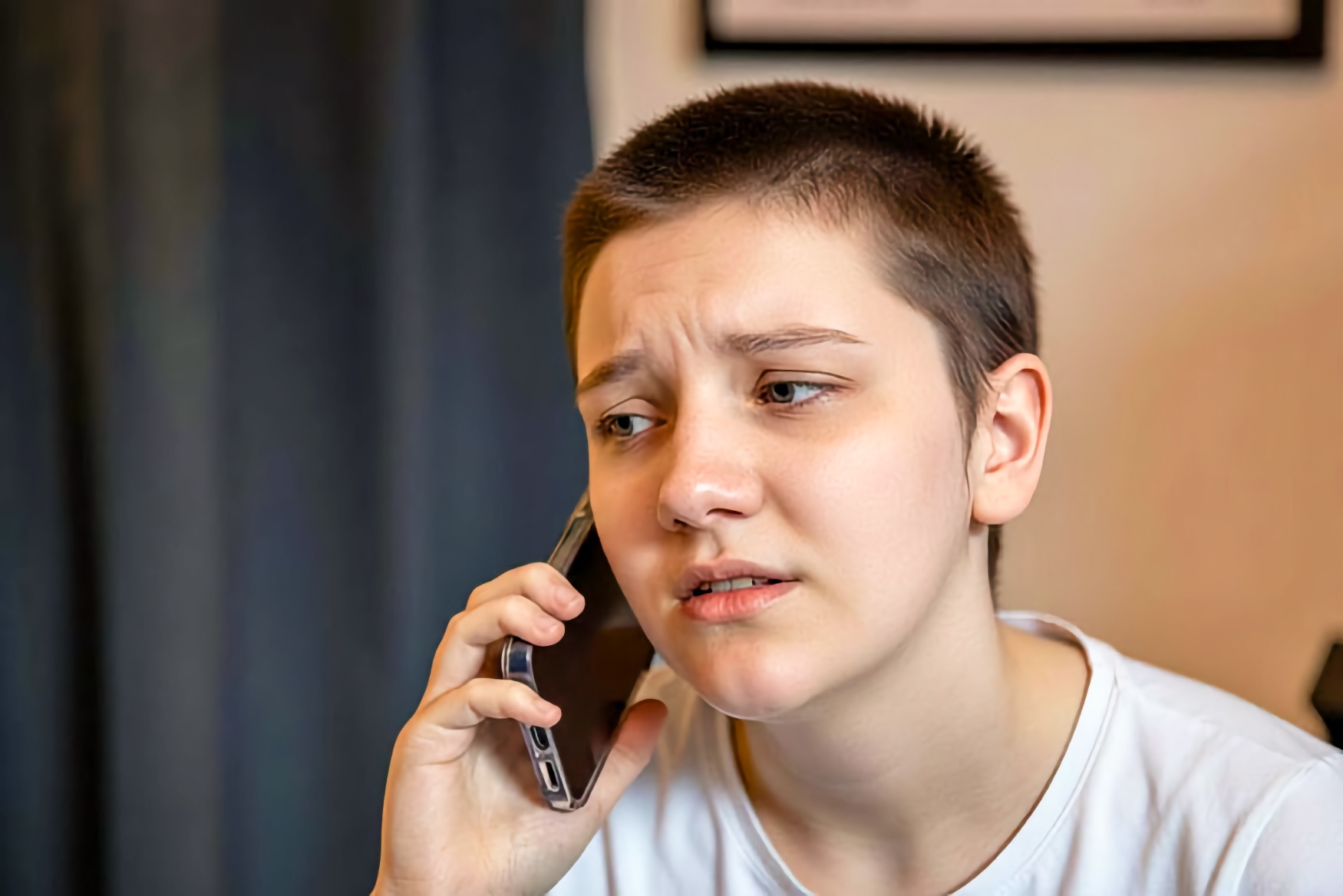 A teenage girl holds a phone to her ear with a worried expression, brow drawn, in a room with a curtain and a framed picture behind her.