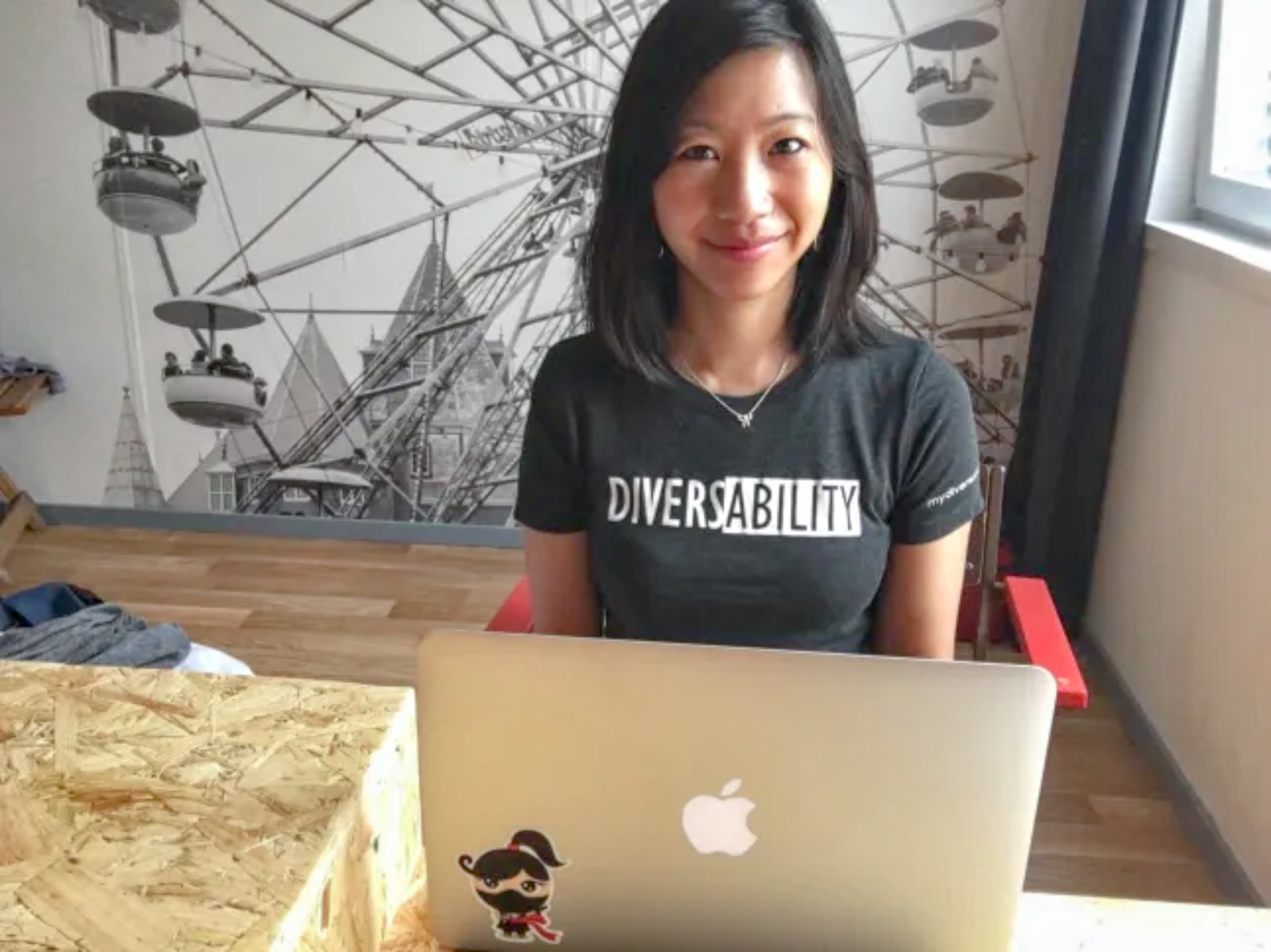 A woman with shoulder-length black hair, wearing a black "DIVERSABILITY" t-shirt, sits at a wooden table with an open silver MacBook that has a ninja sticker. She smiles at the camera. A black-and-white Ferris wheel mural is behind her, and a window to her right lets in natural light.