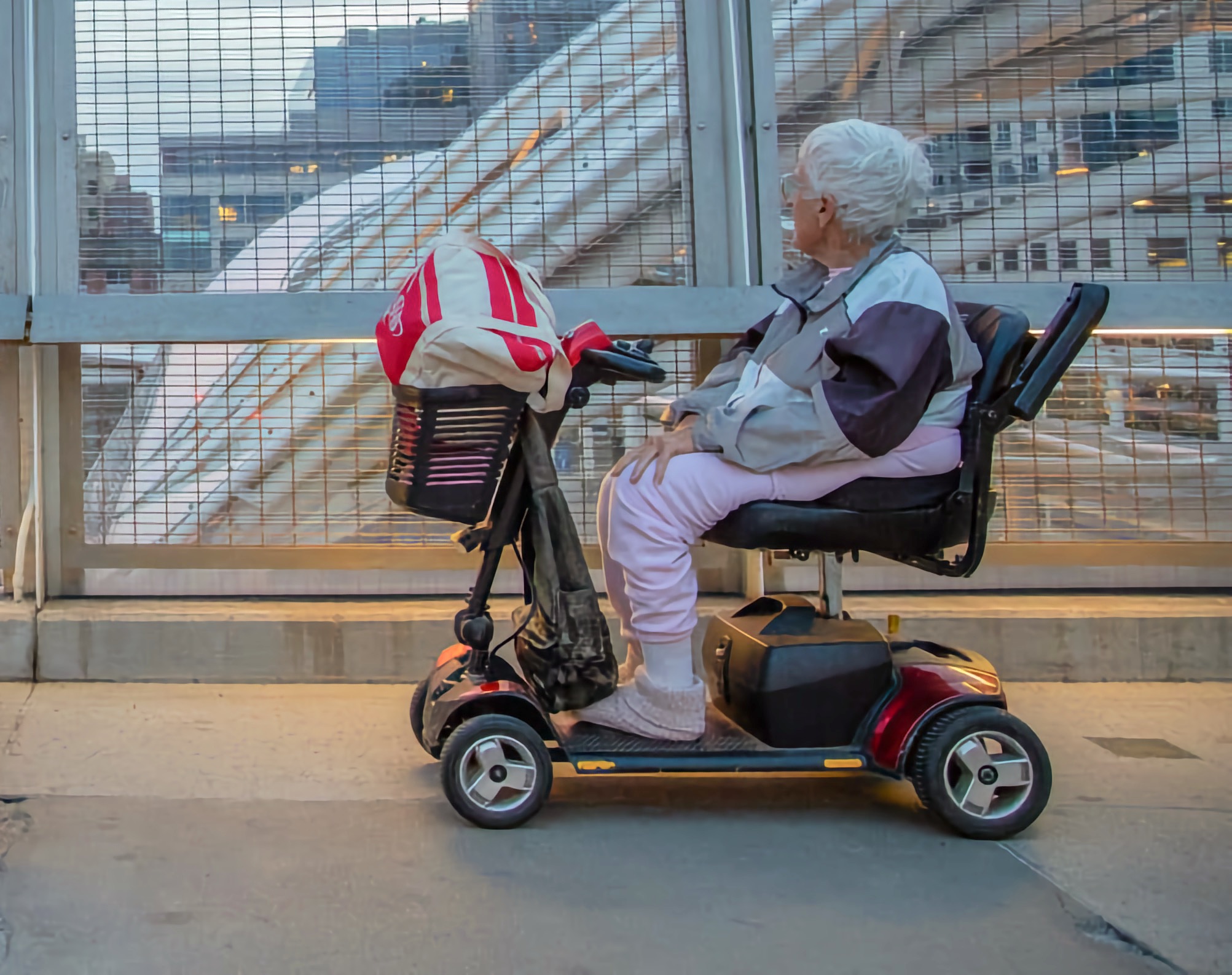 Older woman on a mobility scooter pauses on a city walkway, looking through a wire fence toward the Light Rail lines in Denver.
