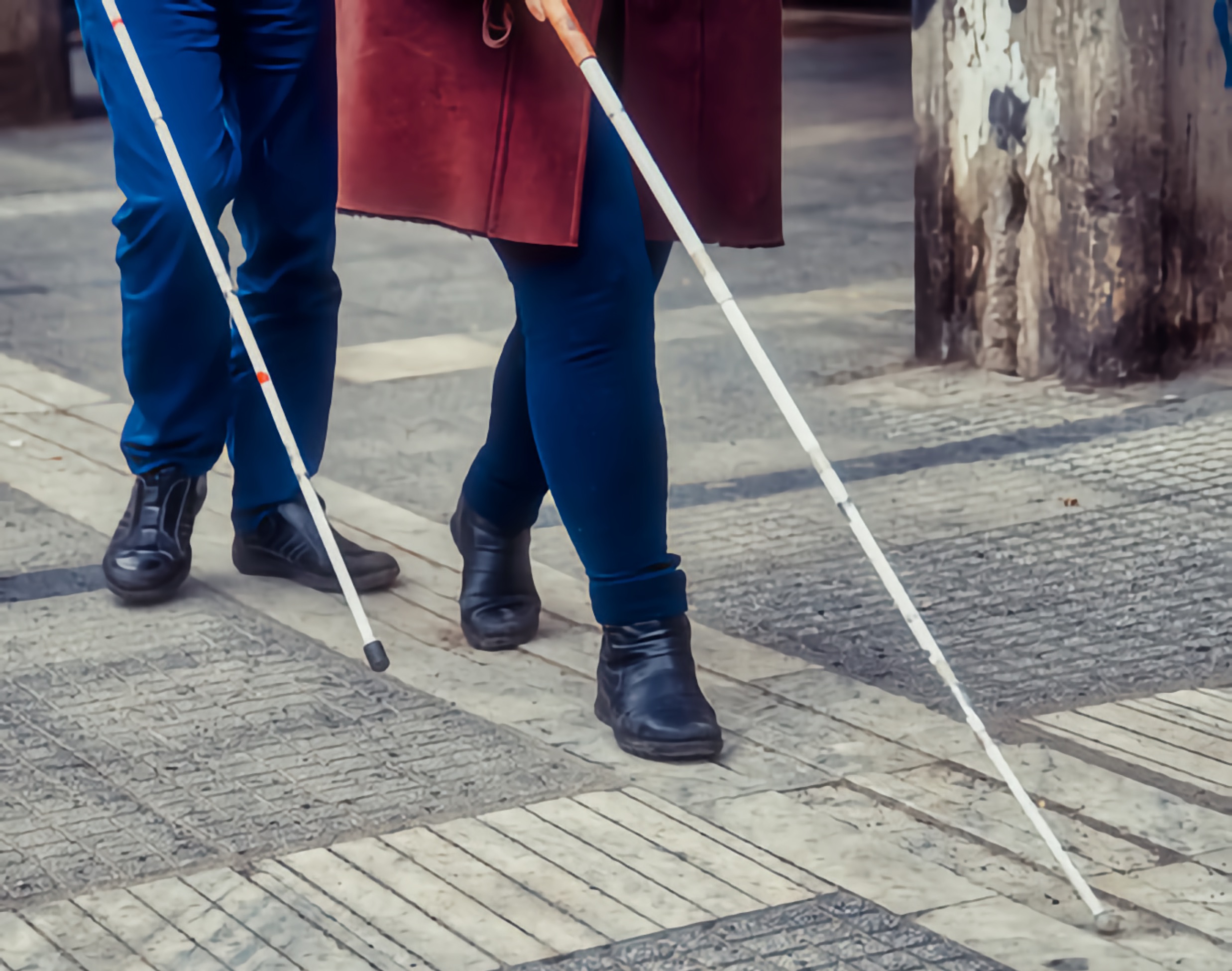 Close-up of two pedestrians using white canes on a textured city sidewalk.