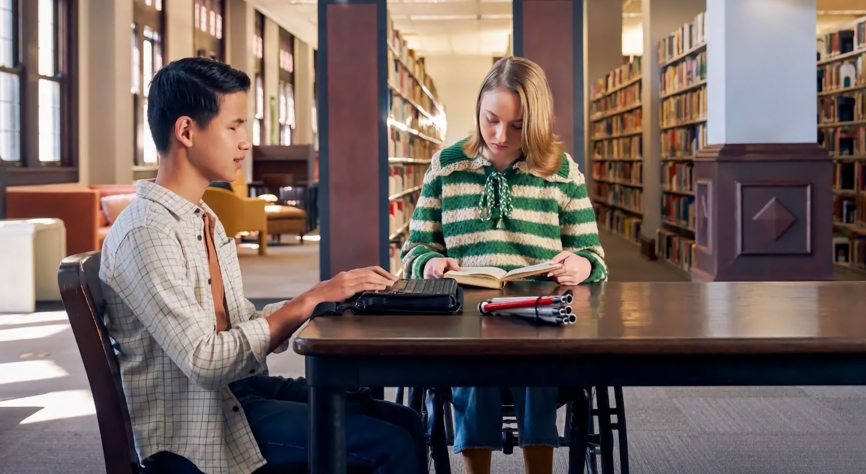 A Blind teenage boy reads a refreshable braille display, his white cane on the table. A teenage girl in a wheelchair reads a book across from him.