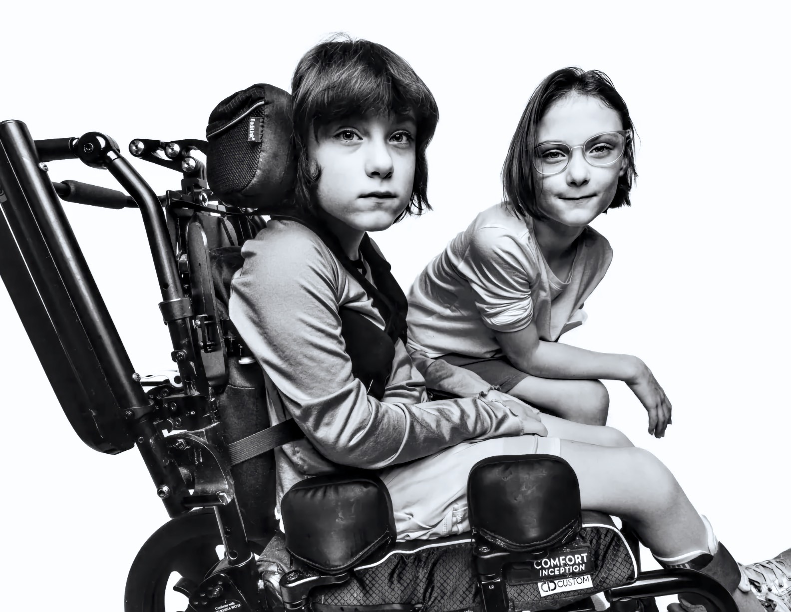 Black and white portrait of two children side by side; one uses a wheelchair with a head support and chest strap, the other wears round glasses.