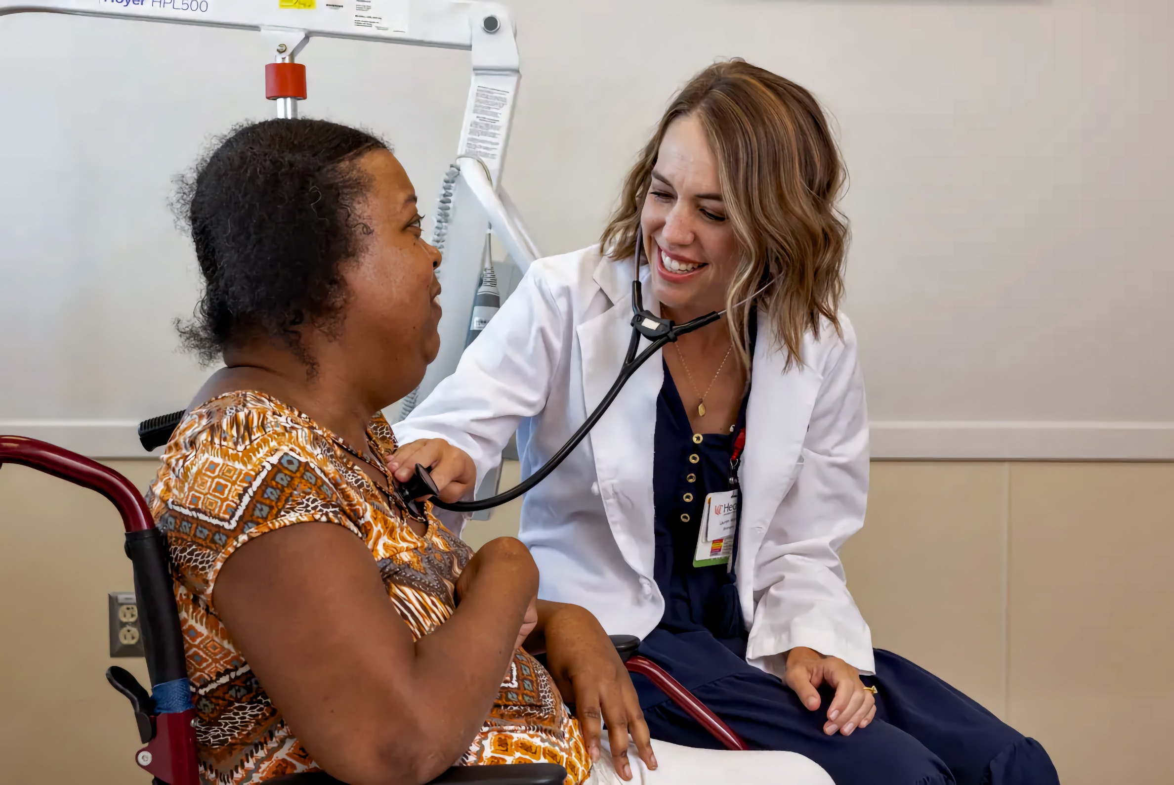 A clinician in a white coat smiles while using a stethoscope to listen to the heart of a middle-aged Black woman seated on a red rollator in a clinic exam room.