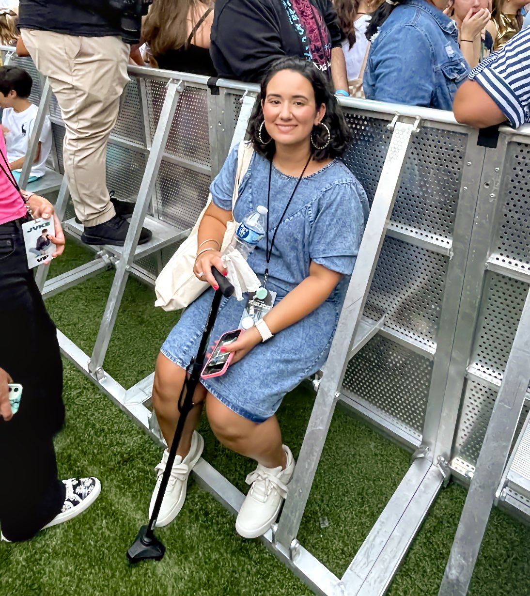 An adult Latina woman sits on a metal bench attached to a barricade at an outdoor concert, holding a cane upright in one hand and a phone in the other, with a standing crowd close around her at the barricade.