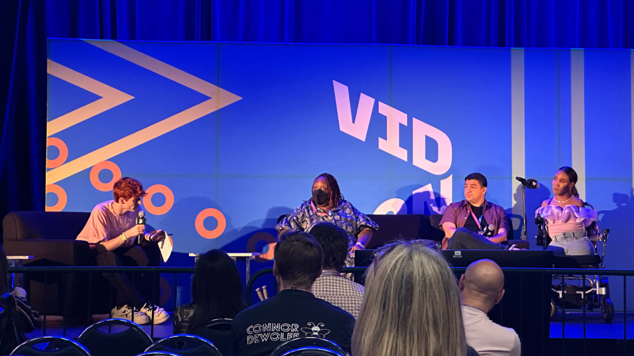 Four people sit on stage under blue lighting with a bold graphic backdrop and microphones in front of them.