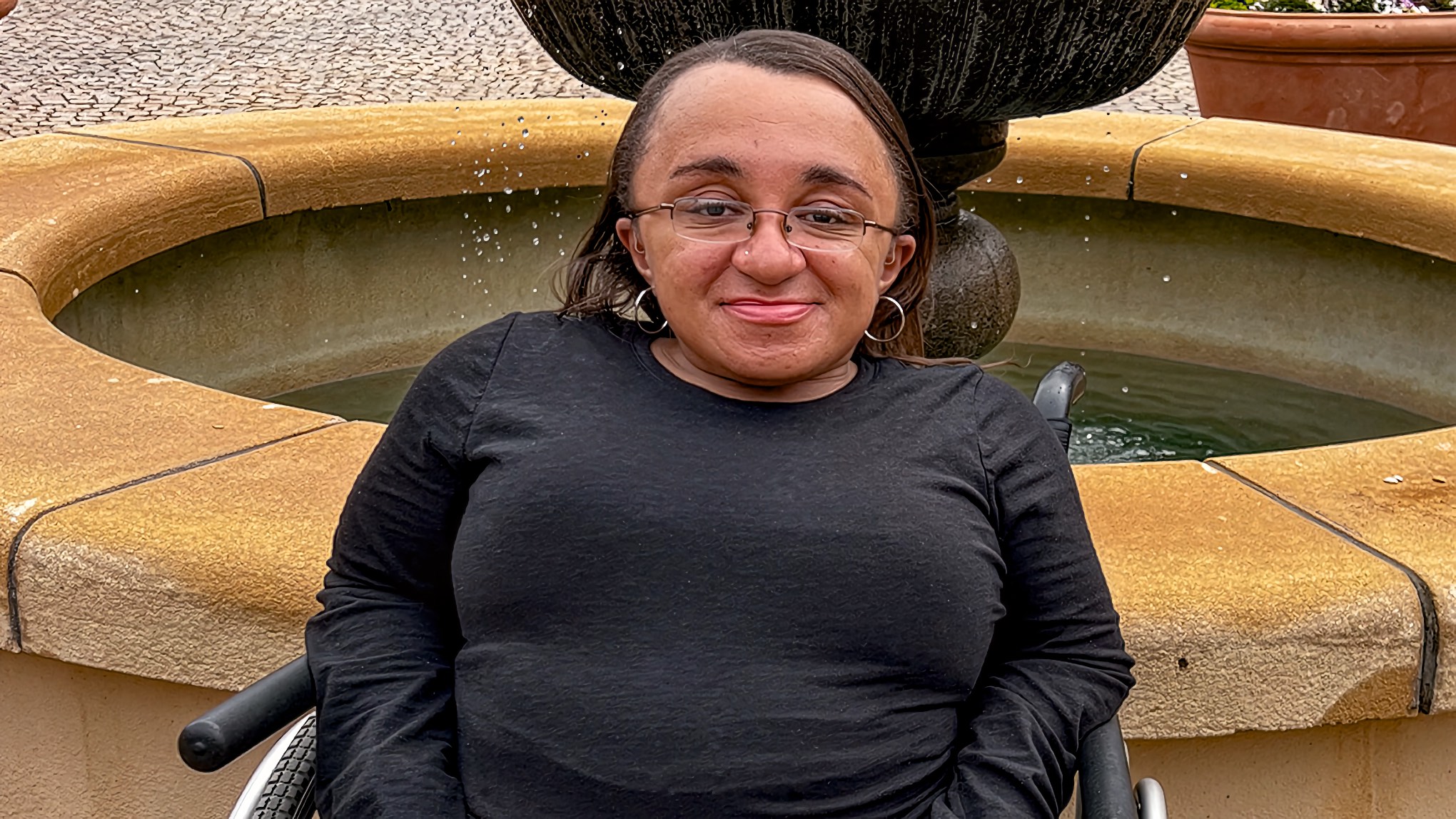 Black woman smiling, seated in a manual wheelchair, wearing glasses, in front of a tiered stone water fountain.