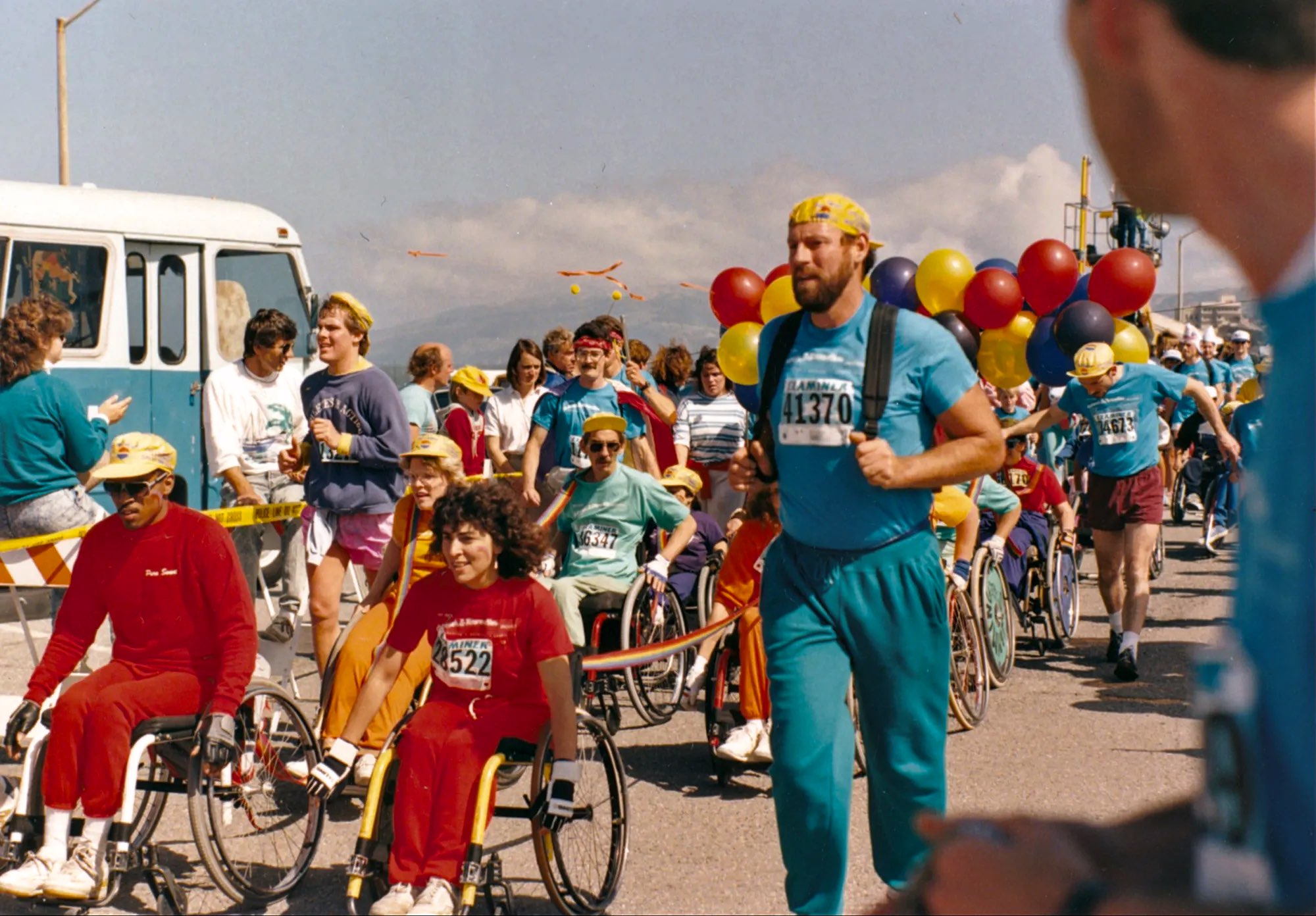 Wheelchair users ride in a linked line down a street during a race, towed by a bearded man wearing a harness, with balloons behind." Long description: A line of wheelchair users, linked one behind another, rolls along a paved street during a road race. A Black man rides at the front left and a woman with dark curly hair rides beside him, both with racing numbers pinned on. A bearded white man wearing a harness and a racing number tows the line on foot, with a bunch of red, yellow, and purple balloons tied behind him. Other wheelchair users and participants on foot follow along the street, a bus is parked at the left, and hills rise in the distance under a clear sky.