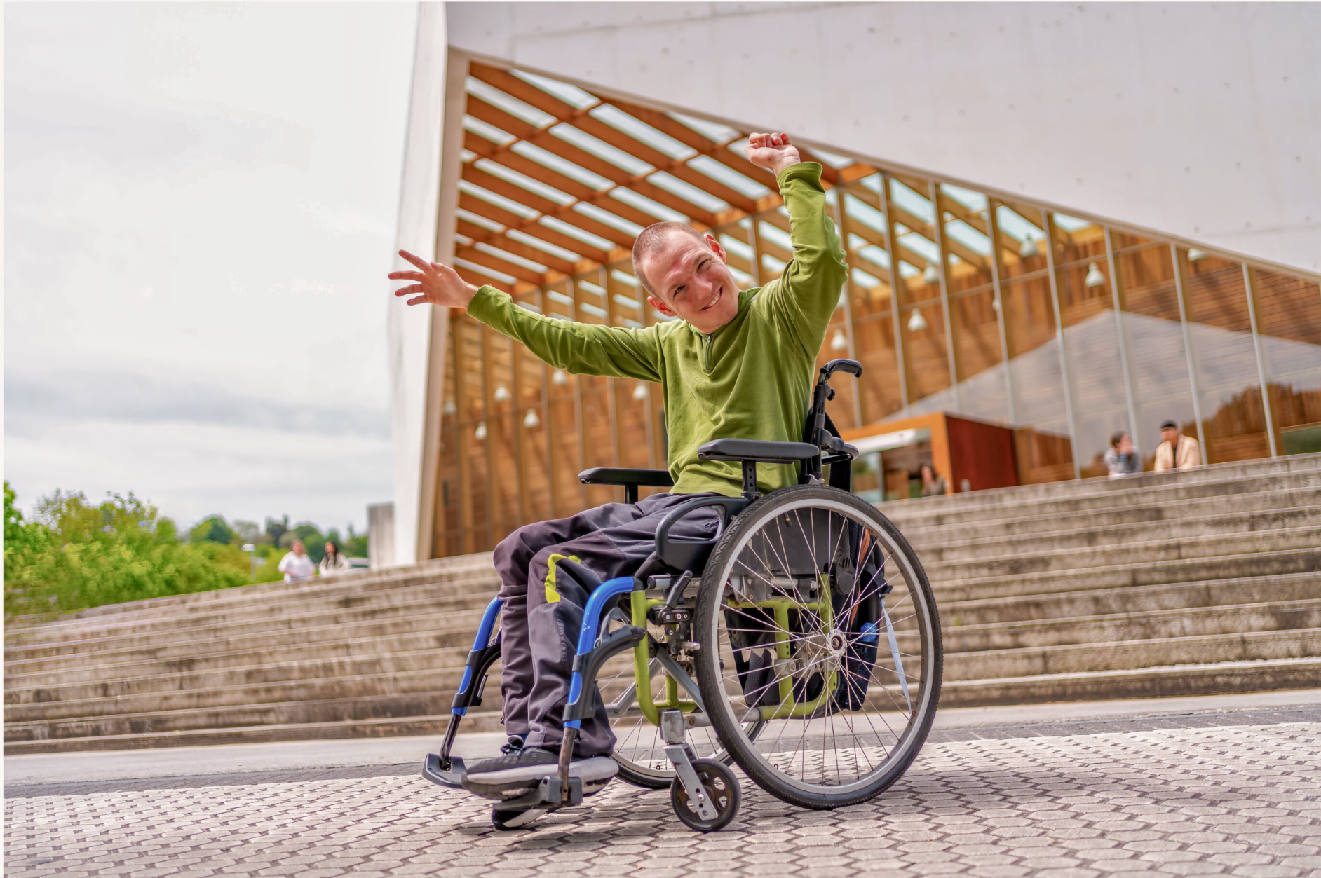 A young adult man, a wheelchair user, raises both arms overhead with hands spread wide and smiles broadly, seated in a green and blue manual wheelchair on a tactile-paving outdoor plaza. Wide concrete stairs and a building with a wood-lattice glass facade are visible behind him.