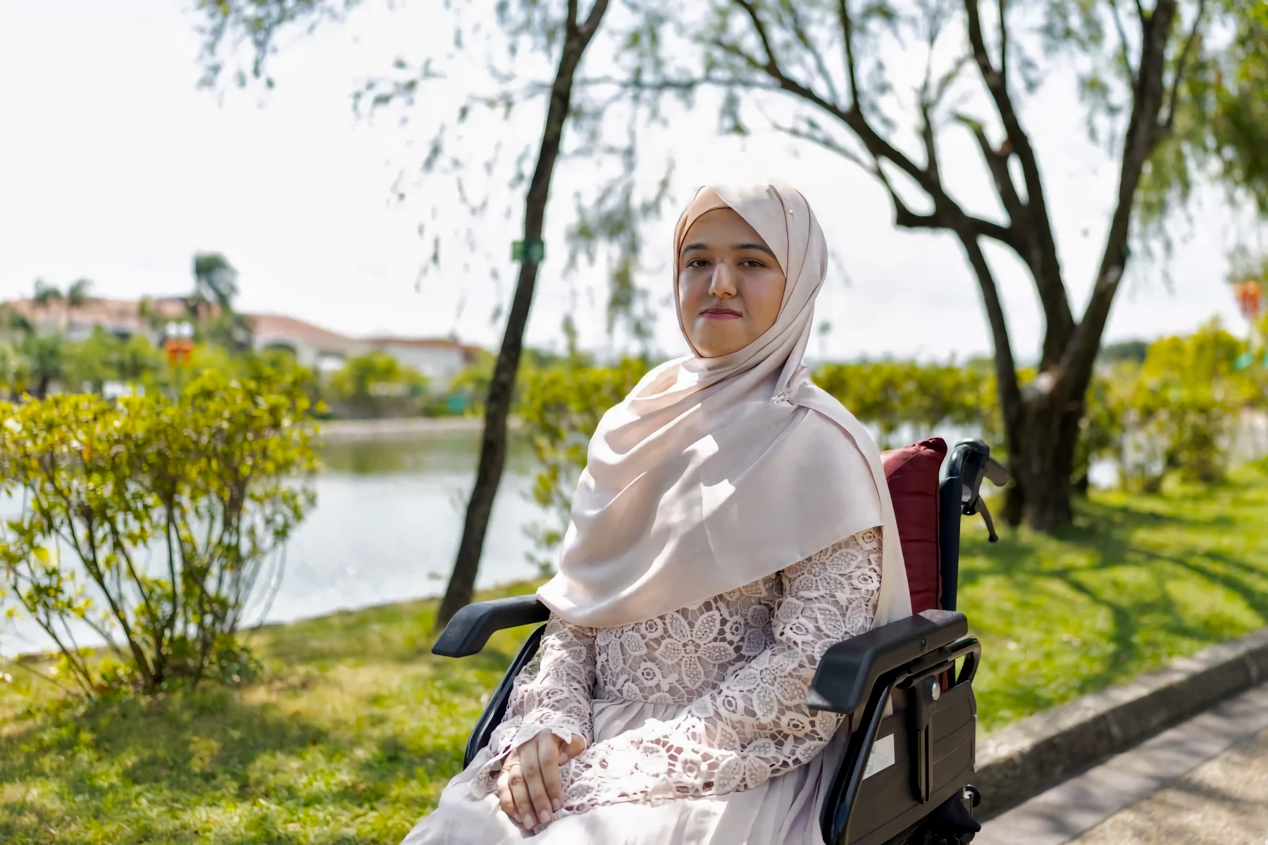 Sa’diyya Nesar in a light hijab in a powered wheelchair, seated by a park lakeside path, looking toward the camera.