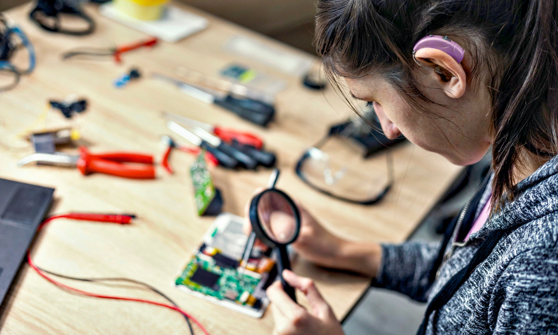 Young woman with a behind-the-ear hearing aid studies a tablet’s open circuit board under a handheld magnifying glass at a cluttered electronics workbench; tweezers in her right hand; safety glasses and soldering tools nearby.