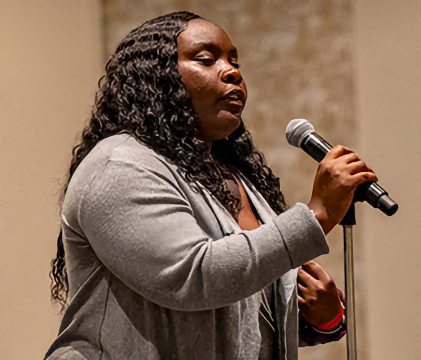 A Black woman with long curly hair wearing a gray cardigan stands at a microphone, her hand gently resting near her chest as she speaks in a softly lit room.