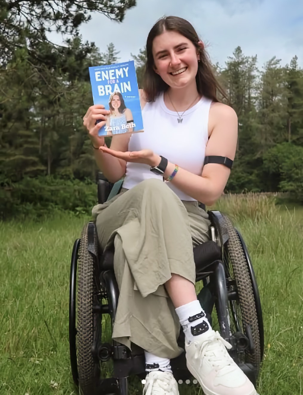 A smiling adult woman seated in a manual wheelchair on a grassy field with trees and sky behind her, holding a book up toward the camera with one hand and her other hand open beneath it. The book cover reads Enemy for a Brain.